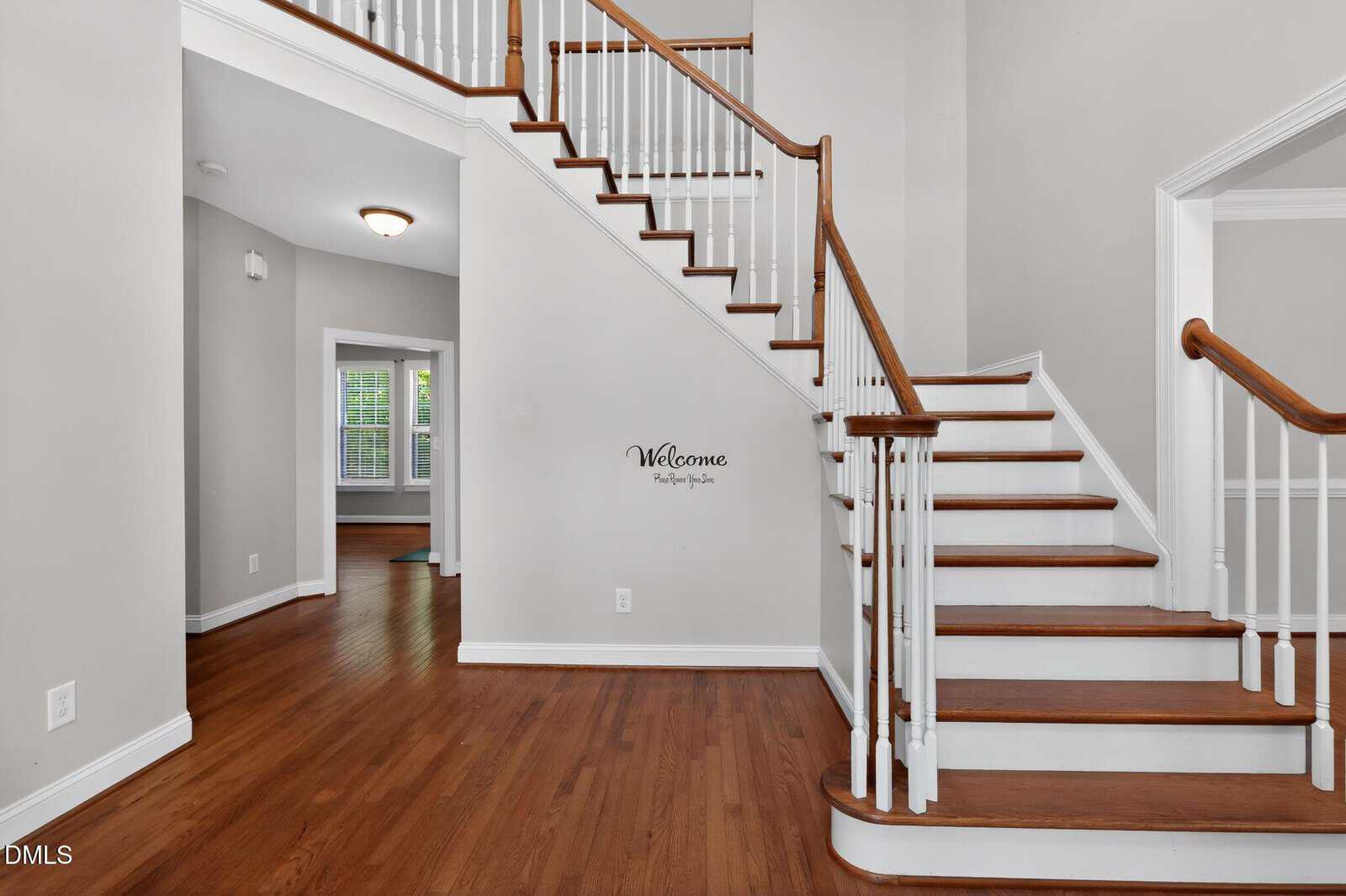 A foyer with wooden floors, white walls, and a staircase with white spindles and wooden handrails. A “Welcome” decal is on the wall, and natural light comes from a window in an adjacent room.