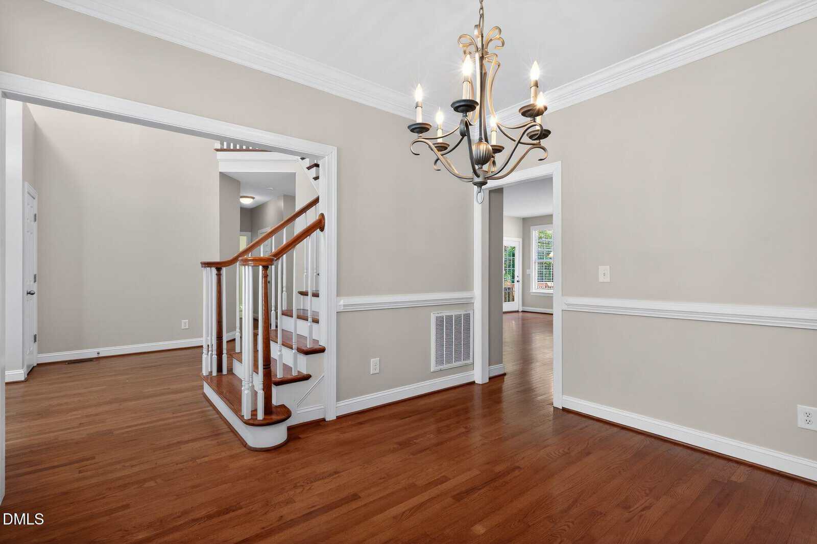 A bright, empty room with hardwood floors, beige walls, white trim, a decorative chandelier, and a wooden staircase with white balusters leading to an upper floor. Multiple doorways open to adjoining rooms.