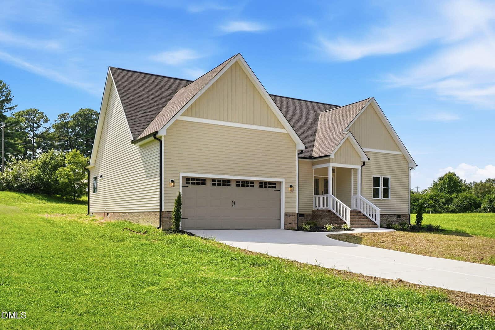Single-story beige house with a two-car garage, gabled roof, front porch, and concrete driveway. The house sits on a grassy lot with trees and open sky in the background.