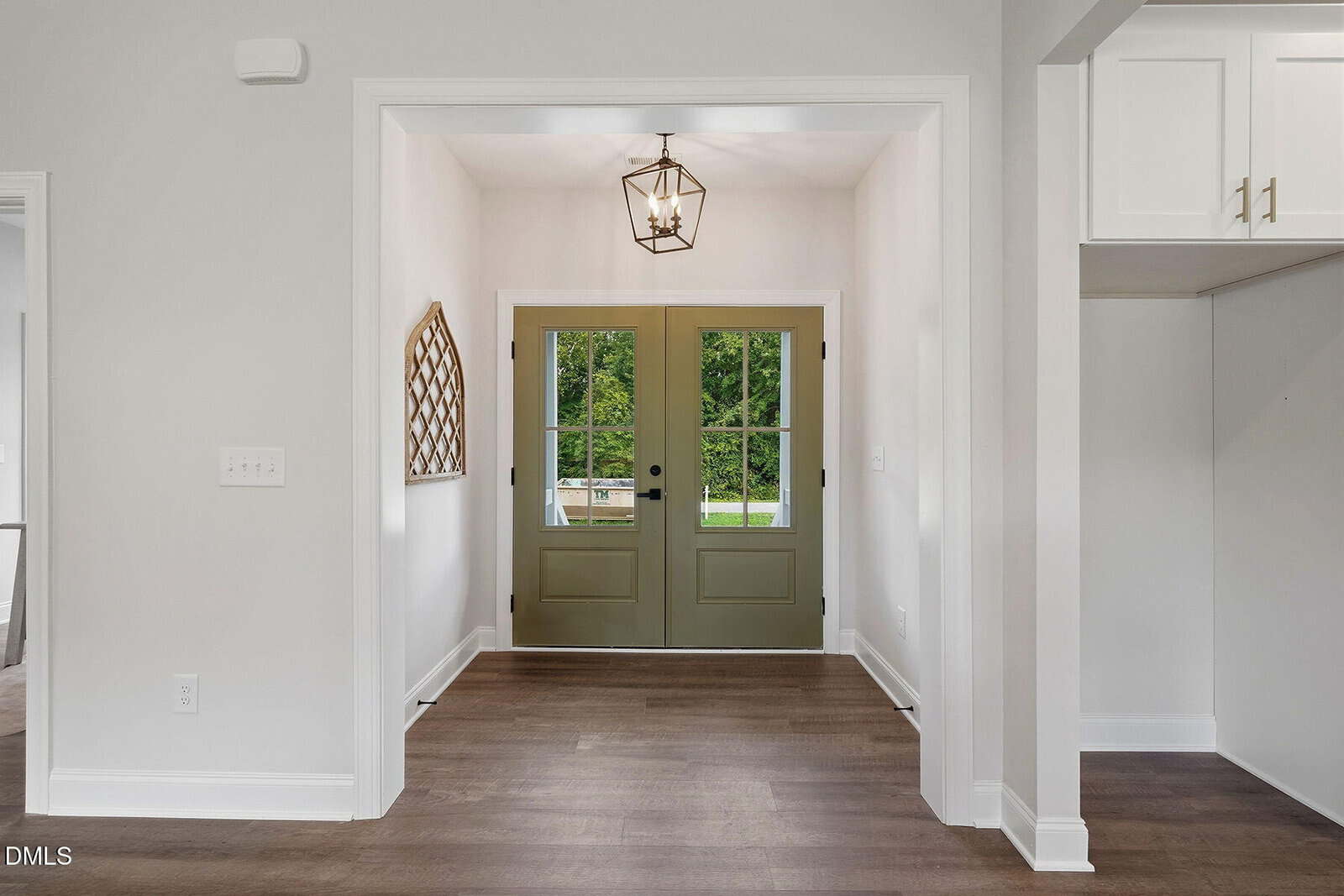 A bright entryway with double olive green doors, a geometric light fixture, wood flooring, white walls, and a decorative mirror on the left wall. A view of greenery is visible through the glass doors.