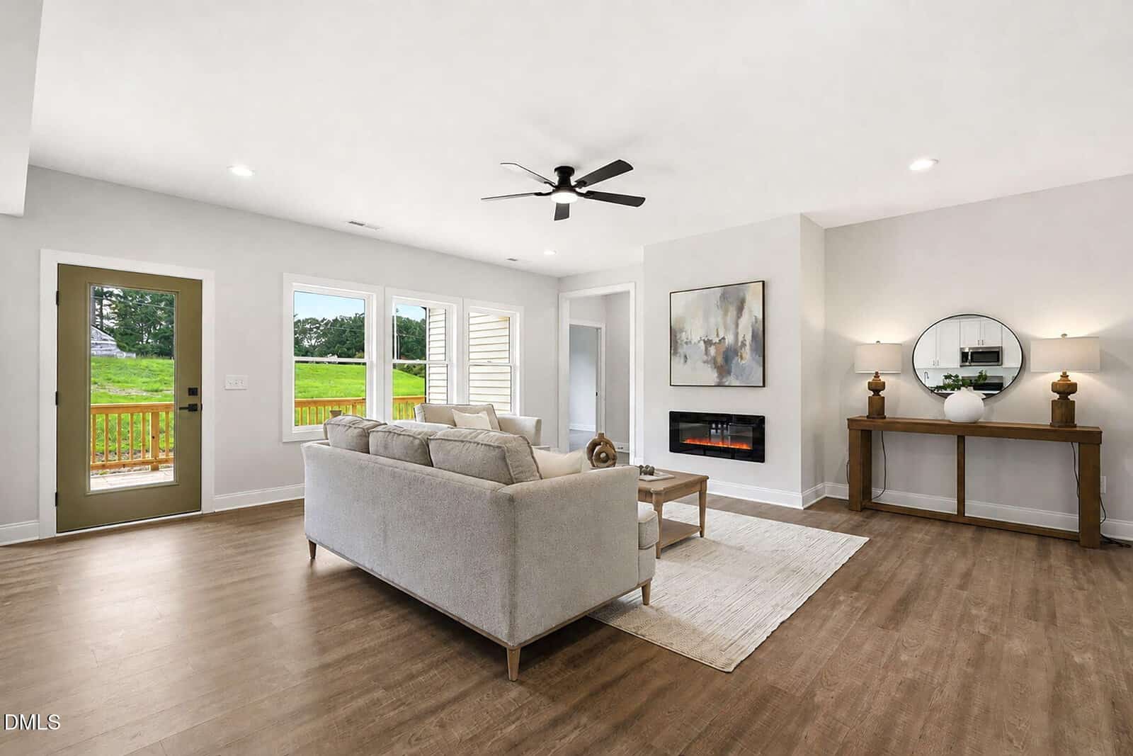 Modern living room with light-colored walls, wood flooring, a beige sofa, a wall-mounted electric fireplace, a console table with decor, large windows, and a glass door leading outside. Ceiling fan and recessed lights are visible.