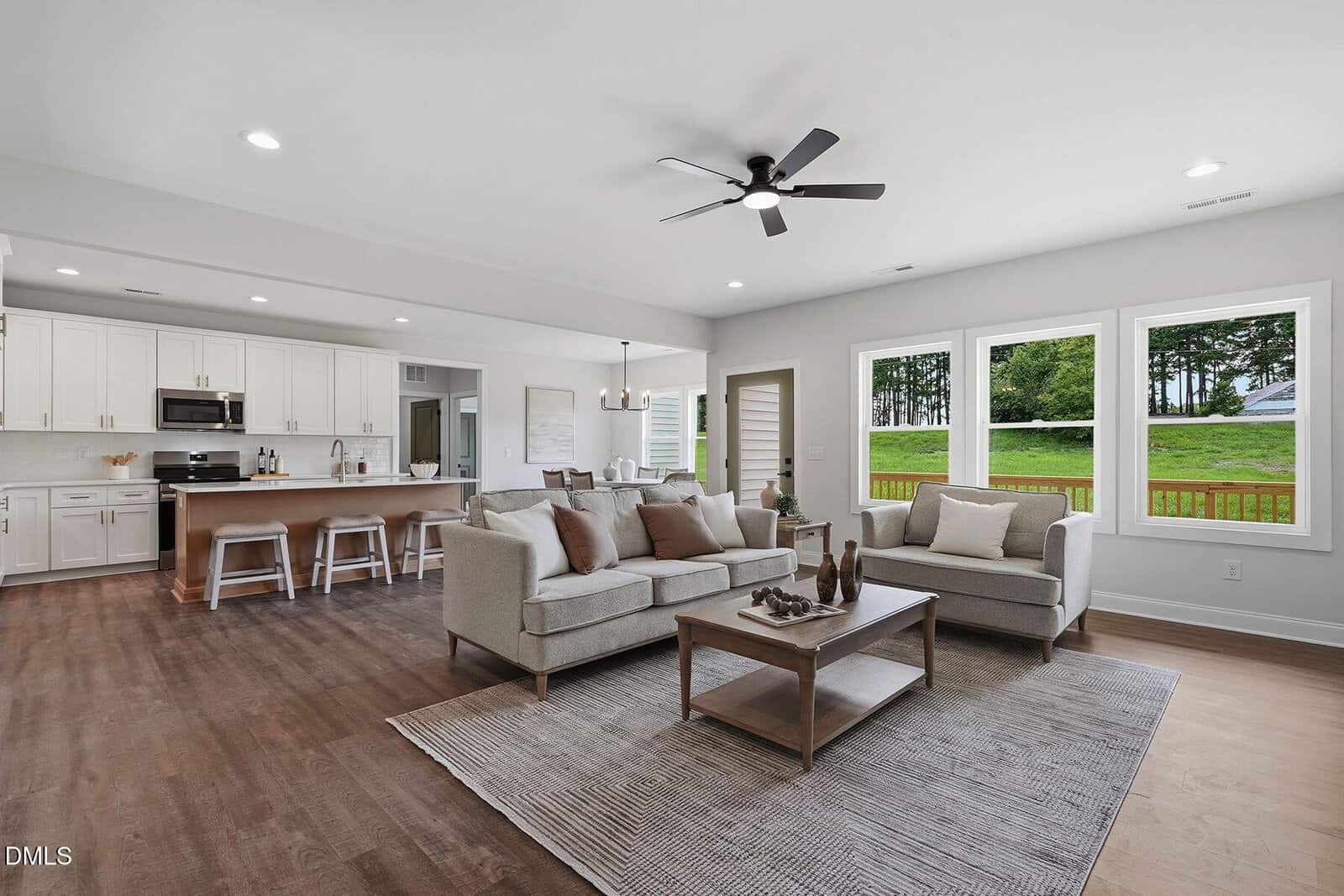Modern open-concept living room and kitchen with neutral decor, large windows showing a green lawn and trees outside, light wood floors, and ceiling fan.