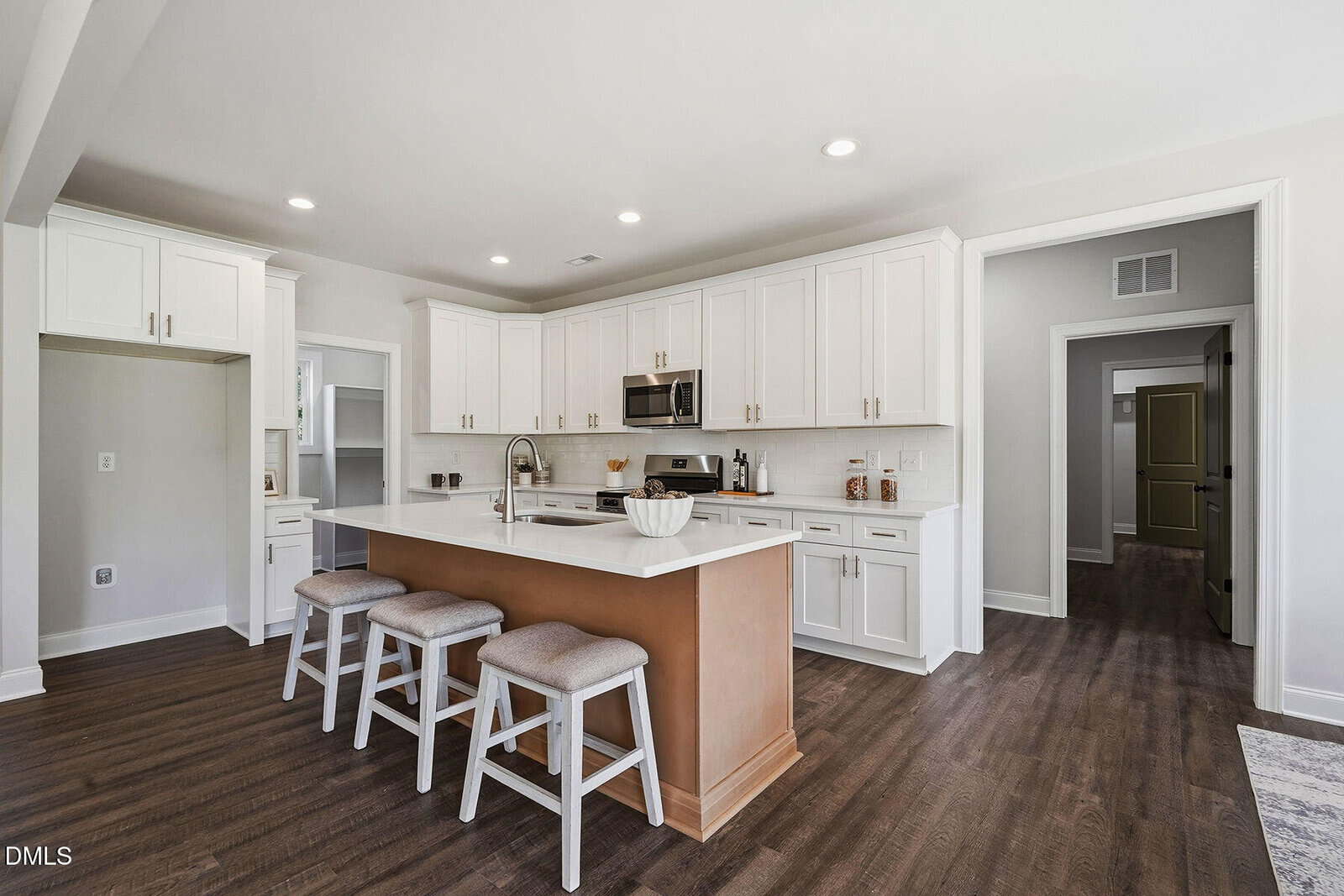 Modern kitchen with white cabinets, stainless steel appliances, a large island with wooden base, three cushioned stools, hardwood floors, and bright lighting. A hallway and doors are visible in the background.
