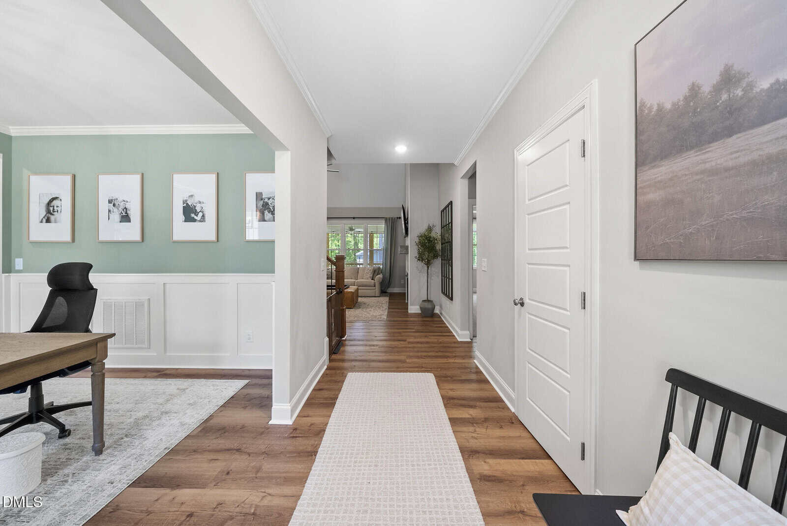 A bright hallway in a modern home with wood floors, white walls, a long runner rug, and a bench. An open office with green accent walls and framed photos is on the left; a living room is visible at the end.