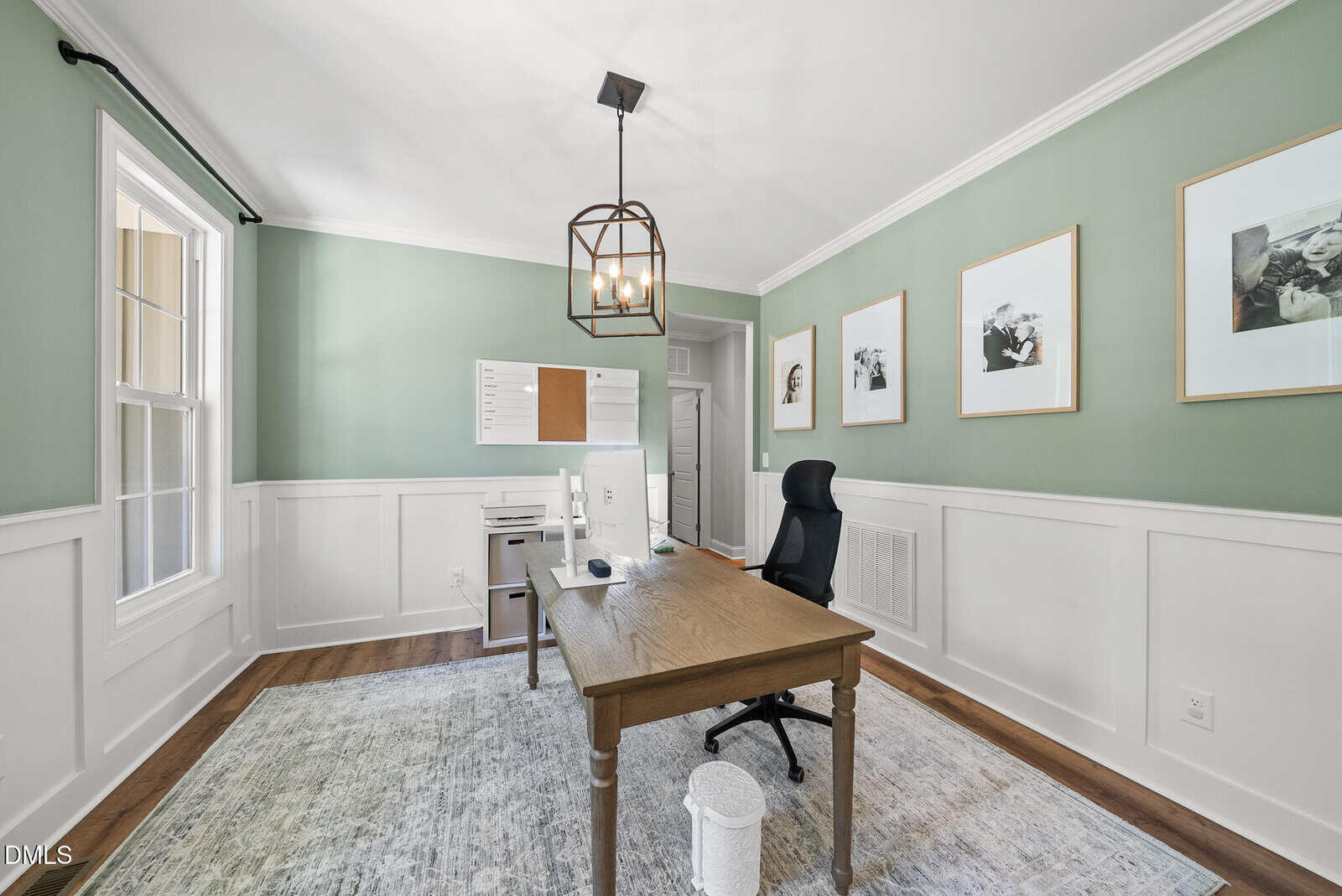 Home office with green walls, wainscoting, wooden desk, black office chair, desktop computer, wall art, rug, and a large window letting in natural light. Modern light fixture hangs from the ceiling.