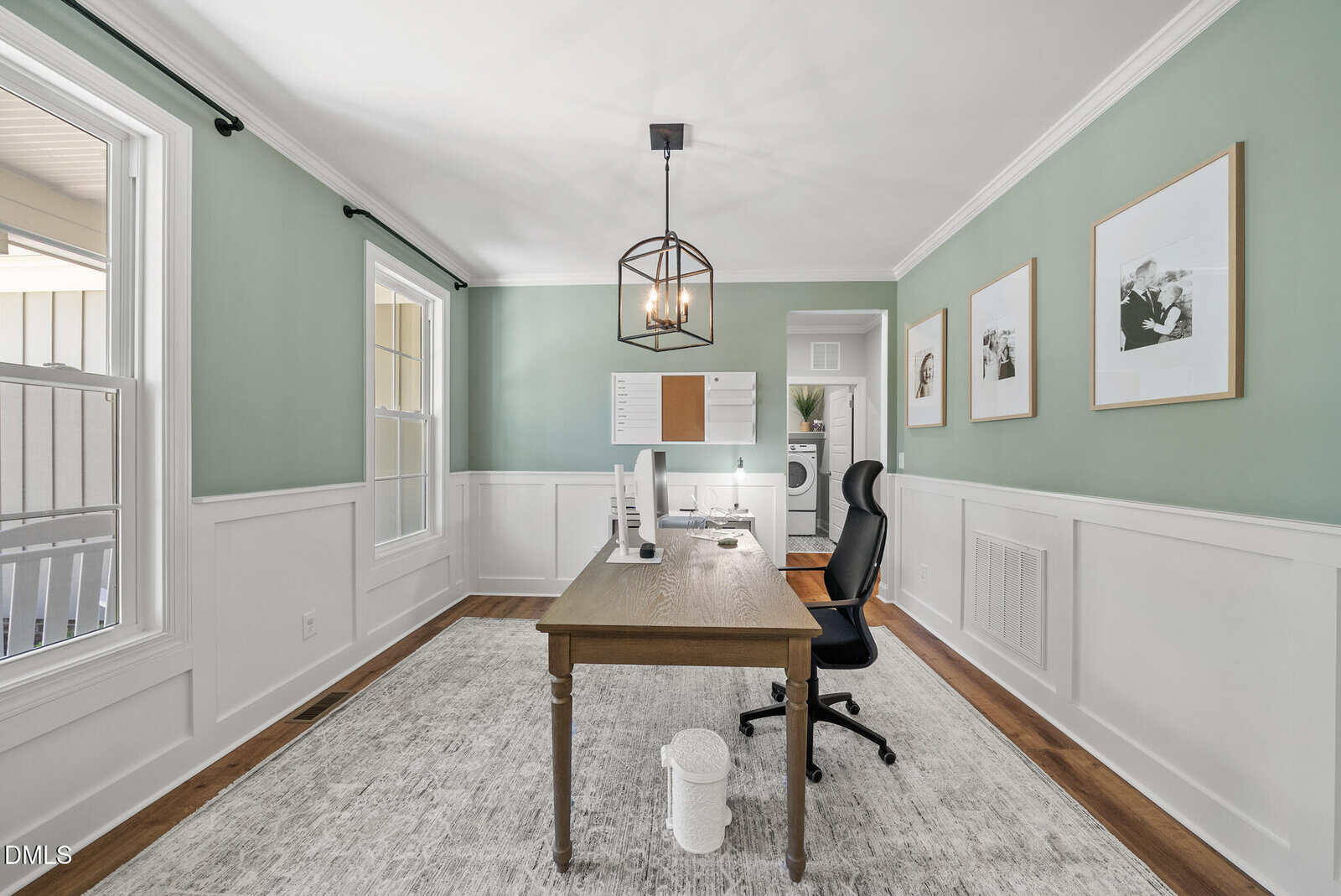 A home office with a wooden desk, black chair, large rug, and green walls with white trim. Wall art, a window, and a board hang above the desk. Laundry appliances are visible through an open doorway.