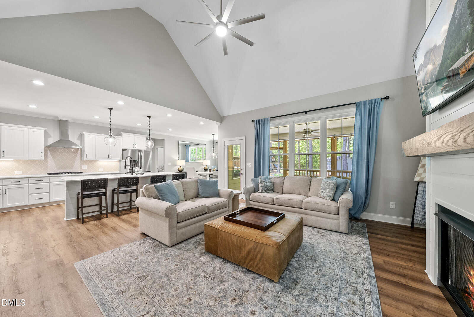 Spacious living room with beige sofas, blue curtains, ottoman, rug, fireplace, and open view into a white kitchen with island seating under recessed lighting and vaulted ceilings. Large windows let in natural light.
