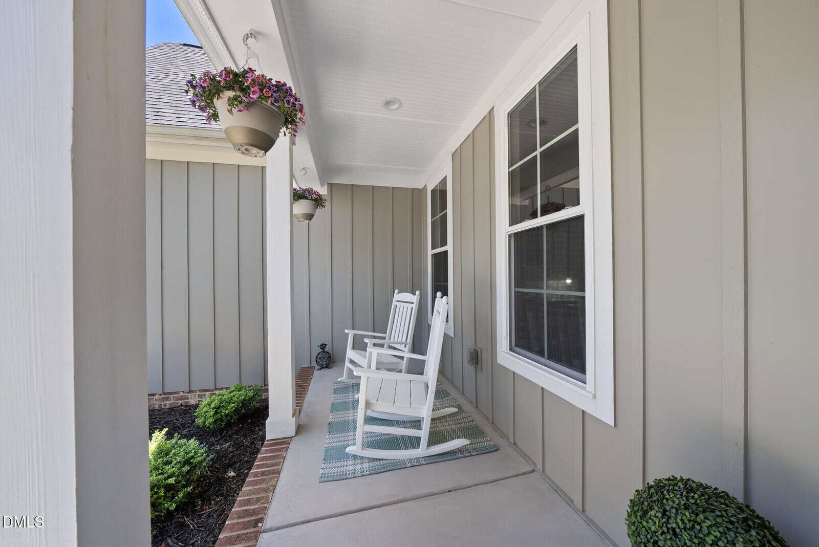 A cozy front porch with two white rocking chairs on striped rugs, hanging flower baskets, and beige vertical siding on the house. Green shrubs line the edge of the porch.