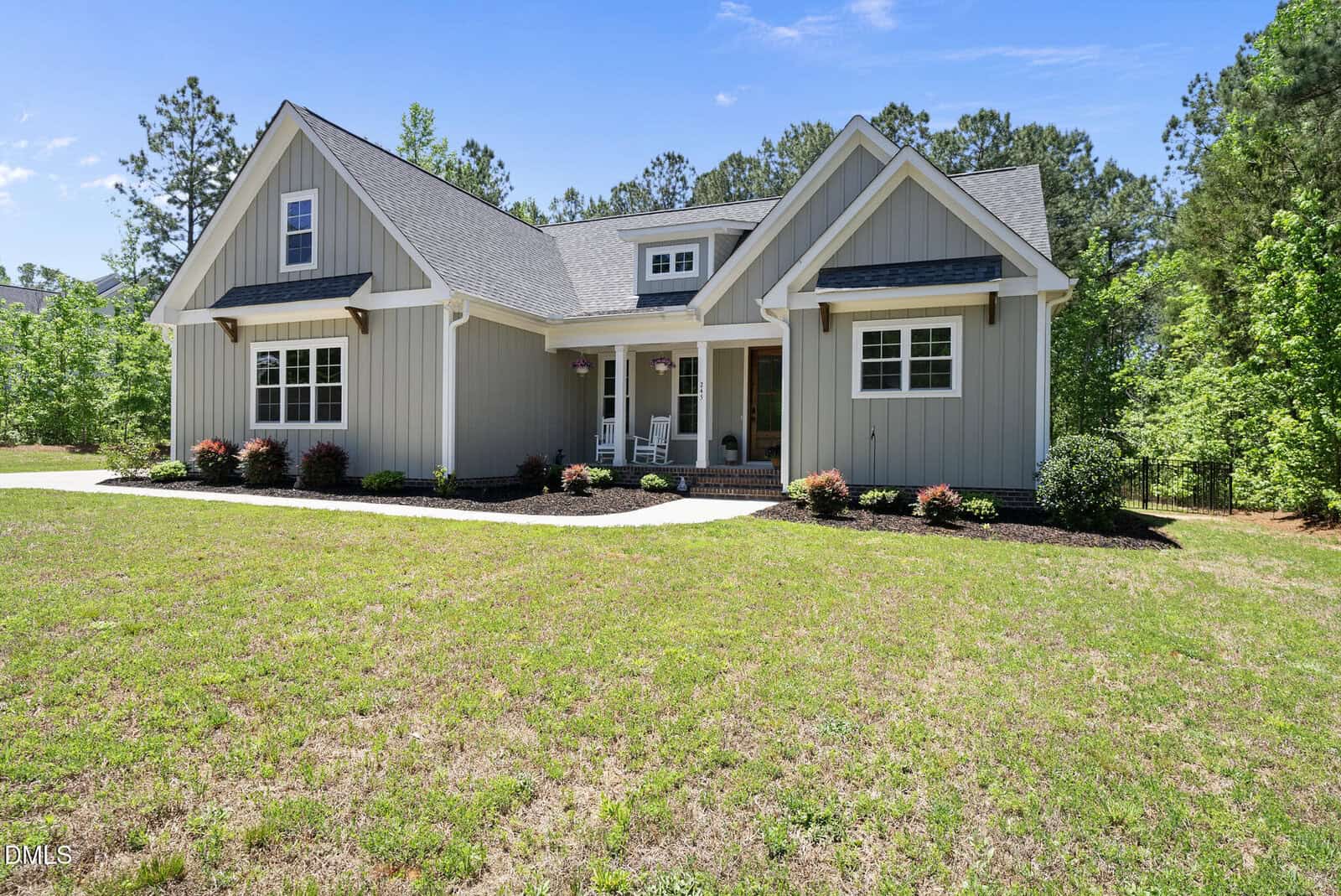 A modern gray house with white trim, a covered front porch with rocking chairs, landscaped flower beds, and a large grassy yard, surrounded by trees under a clear blue sky.