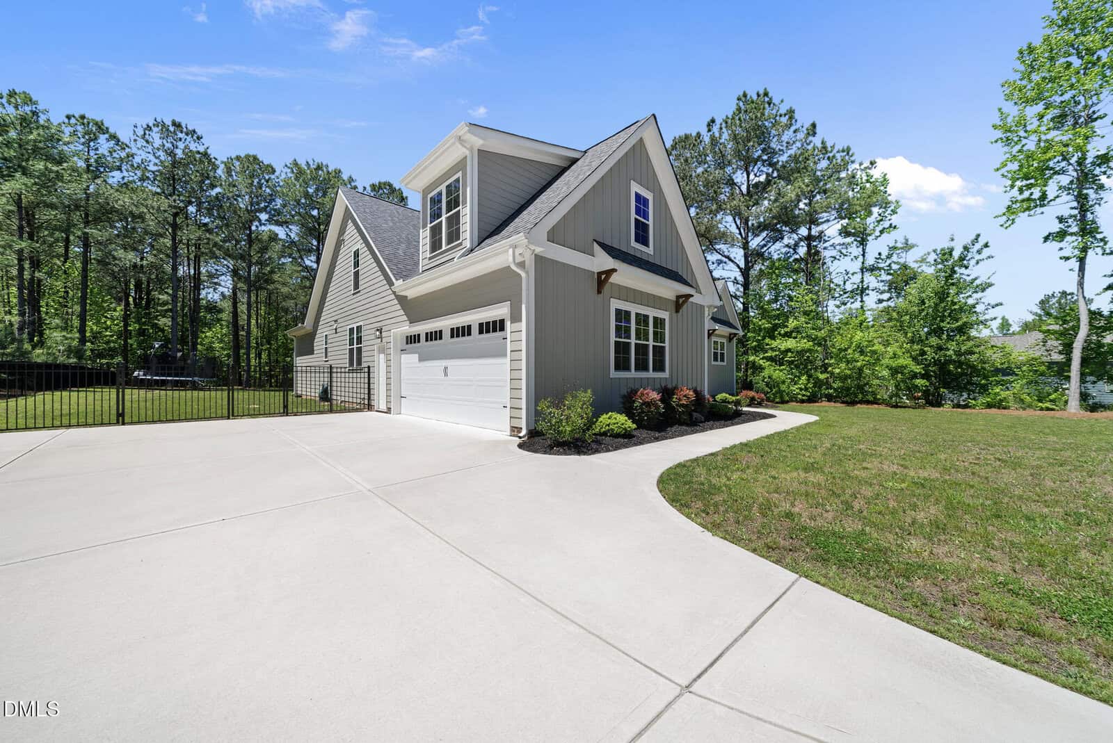 A modern suburban house with gray siding, white trim, and an attached two-car garage sits beside a curved driveway, surrounded by grass, shrubs, and tall trees under a blue sky.