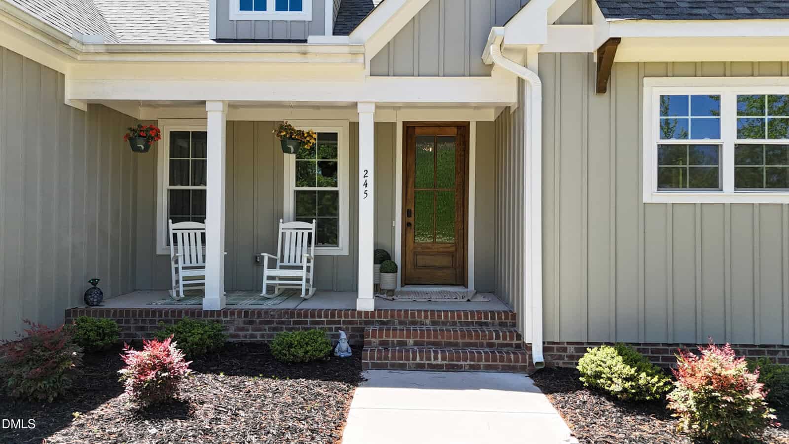 A front porch with two white rocking chairs, hanging flower baskets, a wooden front door with glass panels, and simple landscaping with small shrubs and mulch in front of a gray house.