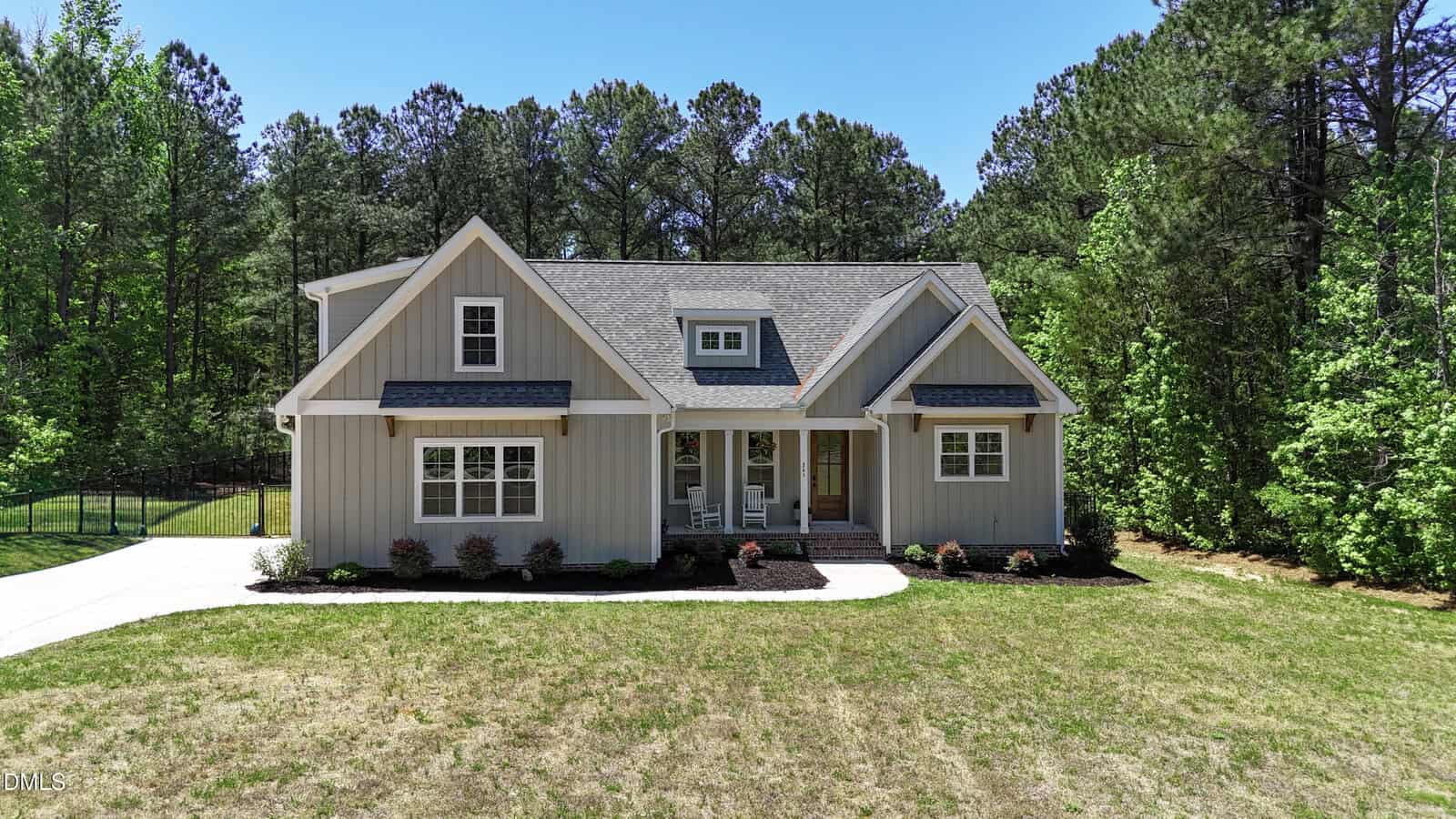 Single-story gray house with white trim and a covered front porch, surrounded by a grassy yard and backed by tall trees under a clear blue sky.
