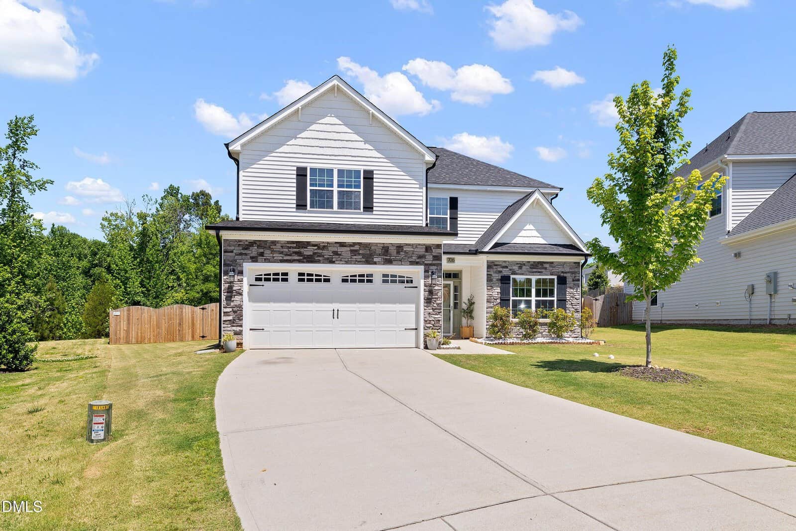 Two-story suburban house with a gray brick and white siding exterior, attached double garage, driveway, small front lawn, and young trees. The sky is blue with scattered clouds.