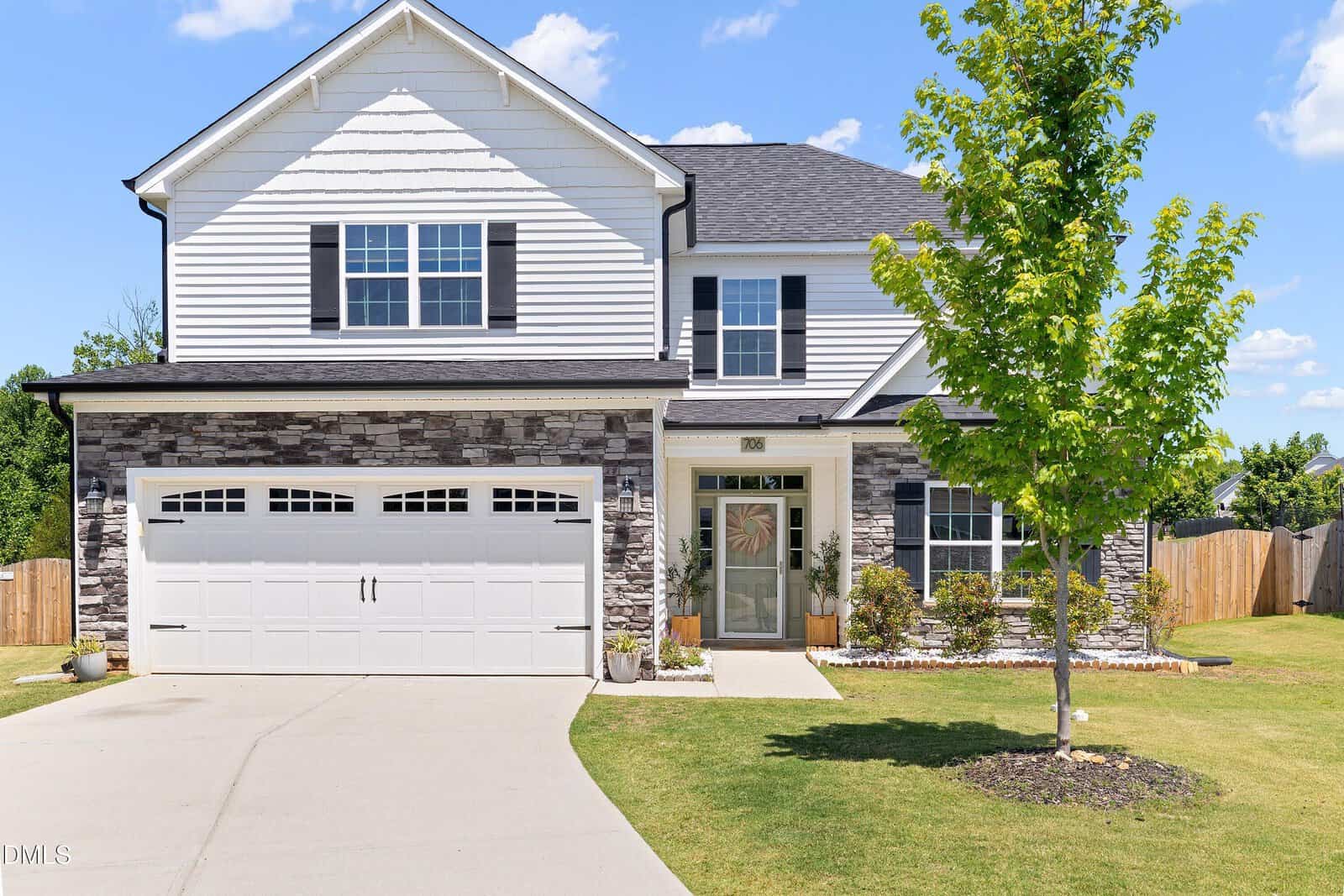Two-story suburban house with white siding, stone accents, a double garage, and a well-kept lawn. A young tree and small shrubs are planted near the entrance. A wooden fence encloses the backyard.