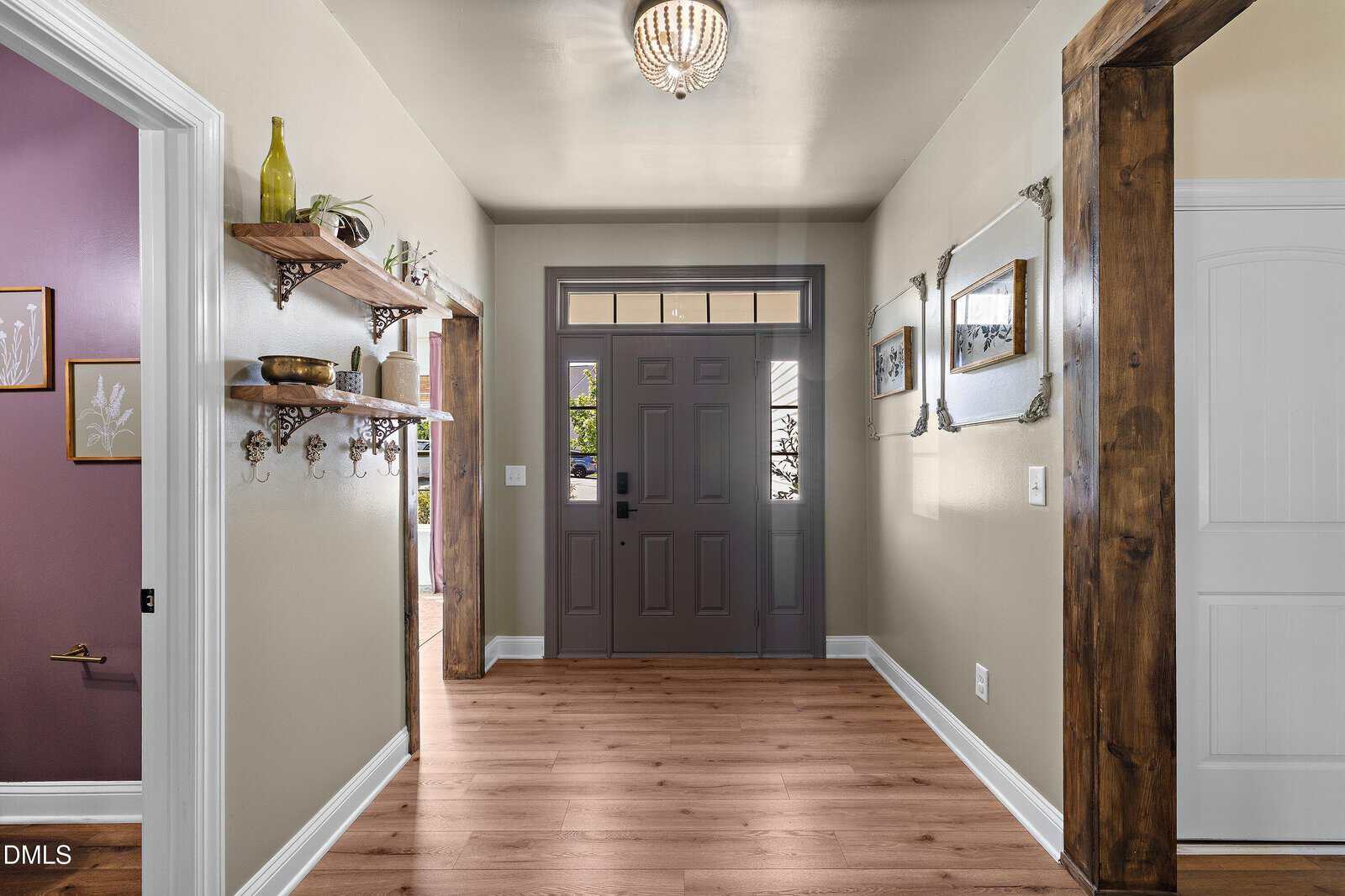 A well-lit entryway features a dark gray front door with glass panels, light wood flooring, neutral walls, wooden shelves with decor, and framed artwork on the right wall. Sunlight streams through the transom window.