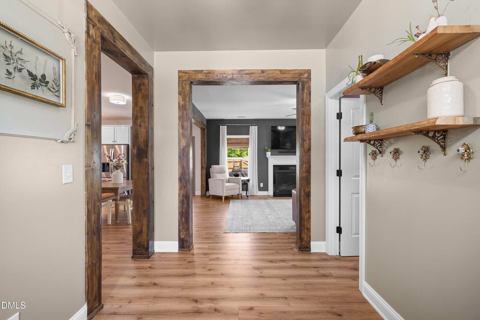 Entryway with wooden floors and rustic wood trims, leading to a living room with a fireplace and mounted TV. To the right, floating shelves hold plants and decor, with hooks and a vase beneath. Dining area visible to the left.