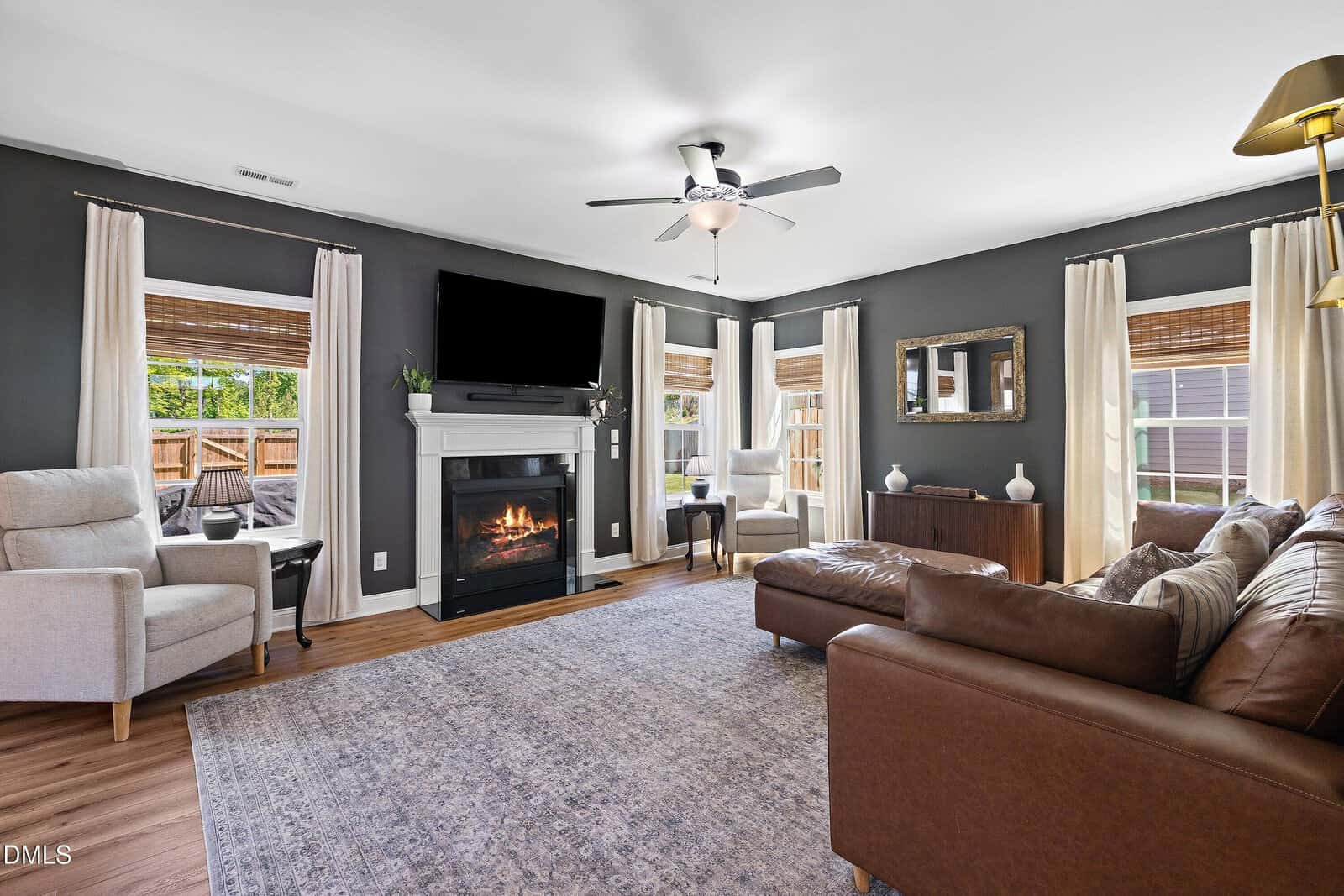 Modern living room with dark walls, a brown leather sofa, gray armchairs, fireplace, wall-mounted TV, large windows with curtains, and a blue patterned rug on wood flooring.
