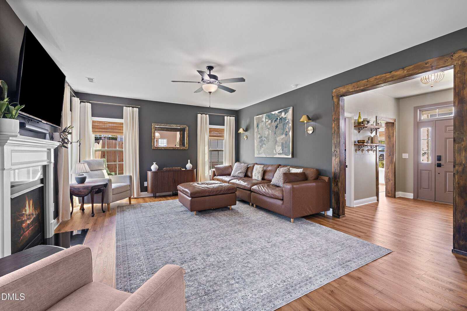Spacious living room with gray walls, wooden floors, brown sectional sofa, large area rug, fireplace with mounted TV, armchairs, artwork, and multiple windows with light curtains letting in natural light.