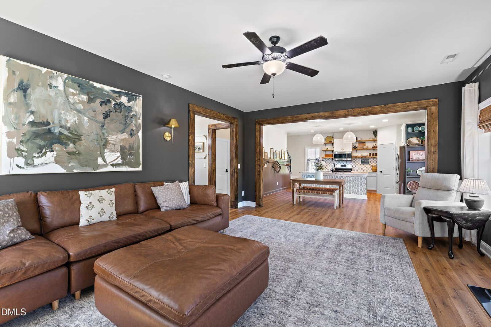 A cozy living room with a brown leather sectional, abstract art on a dark wall, and a ceiling fan. The room opens to a bright kitchen with an island and wooden floors. Large windows provide natural light.