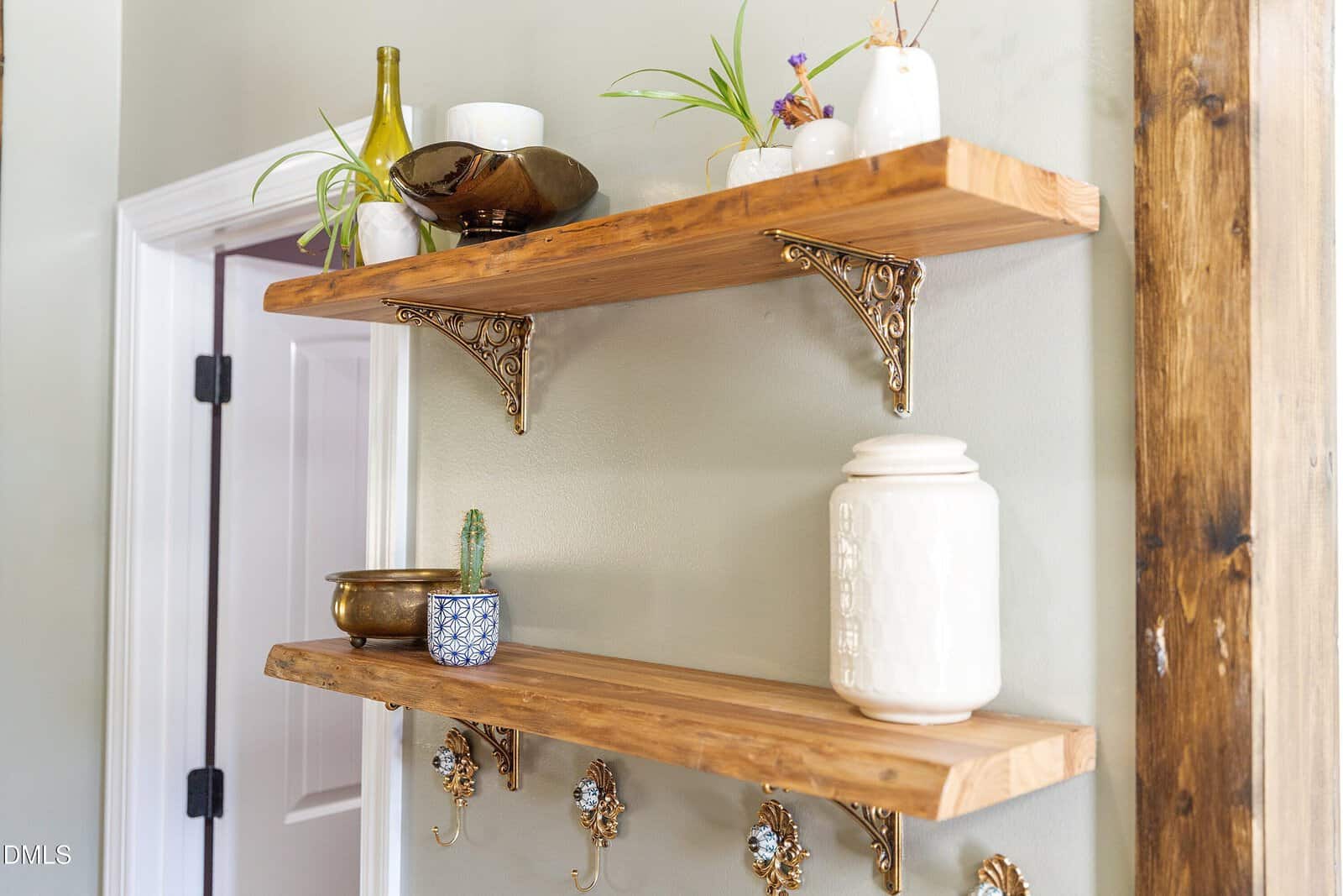 Two wooden wall shelves with ornate gold brackets display potted plants, vases, and decorative bowls. A white ceramic jar sits on the lower shelf. The shelves are mounted on a light-colored wall near a door.