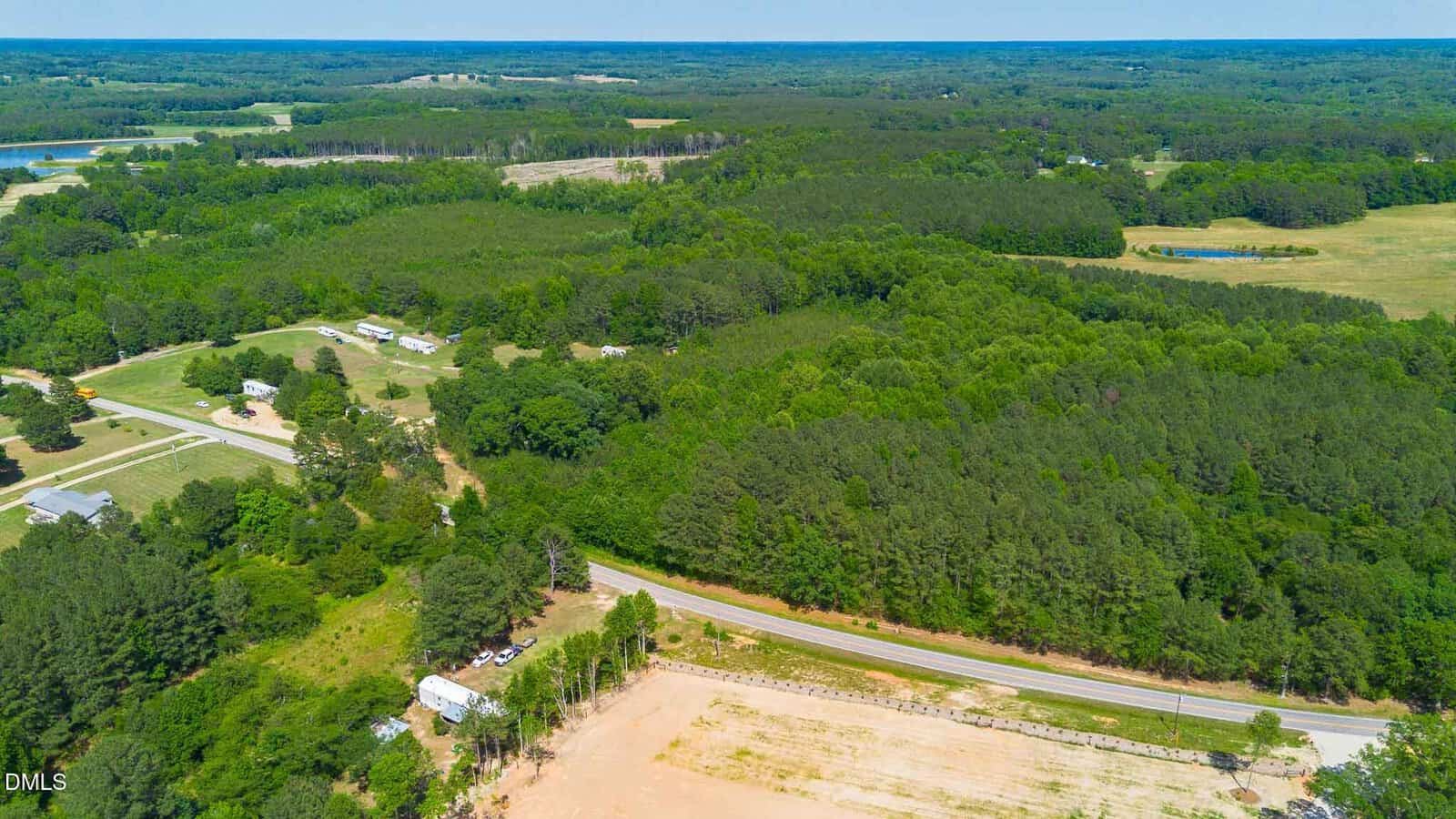 Aerial view of a rural area with dense forest, scattered houses, open fields, and a road curving through the landscape under a clear blue sky.
