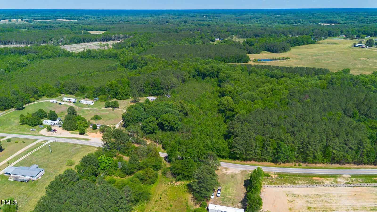 Aerial view of a rural landscape with scattered houses, winding roads, open fields, and dense green forests under a clear blue sky.