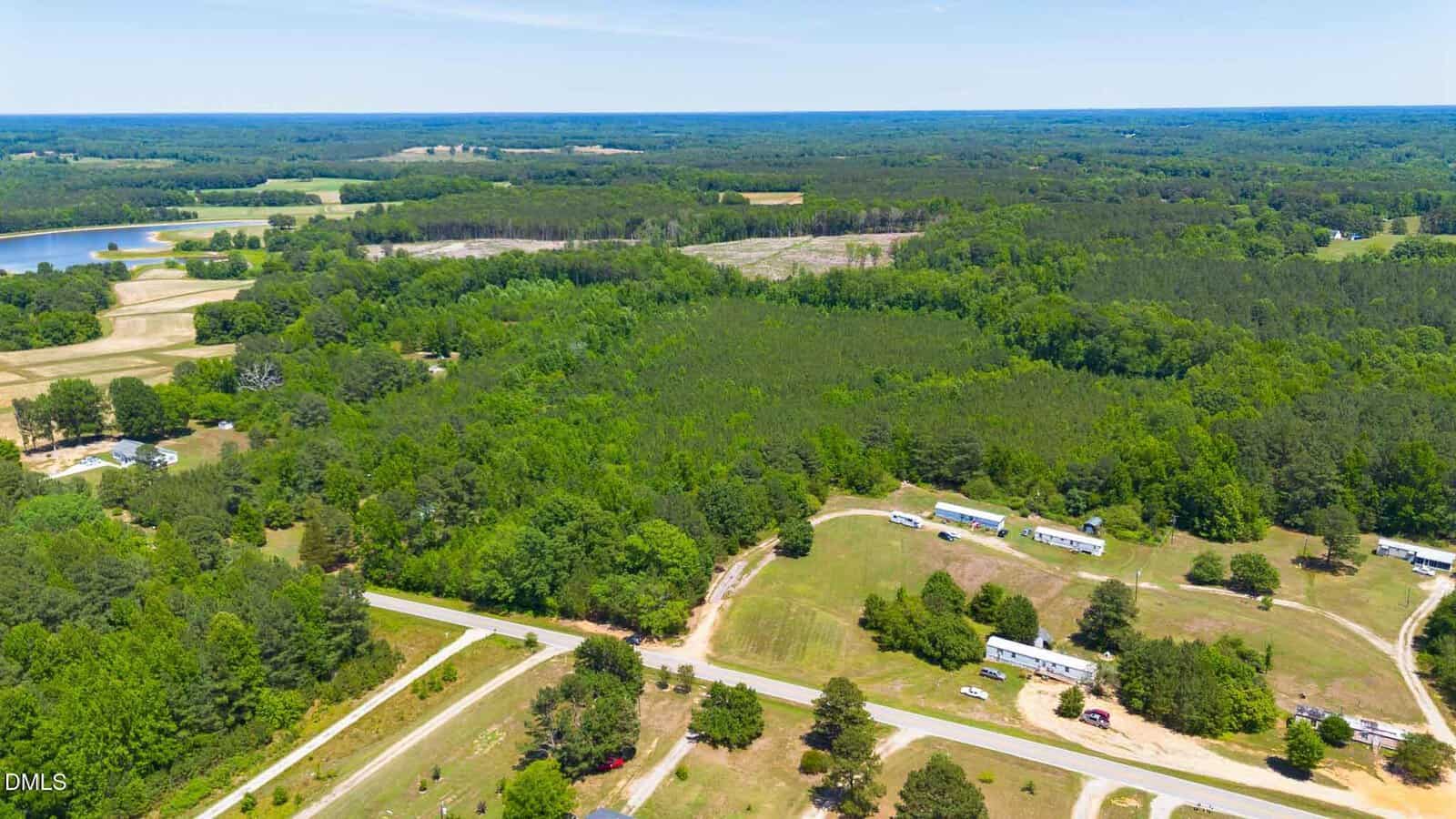 Aerial view of rural landscape with scattered homes, winding roads, dense green forests, open fields, and a pond in the distance under a clear blue sky.