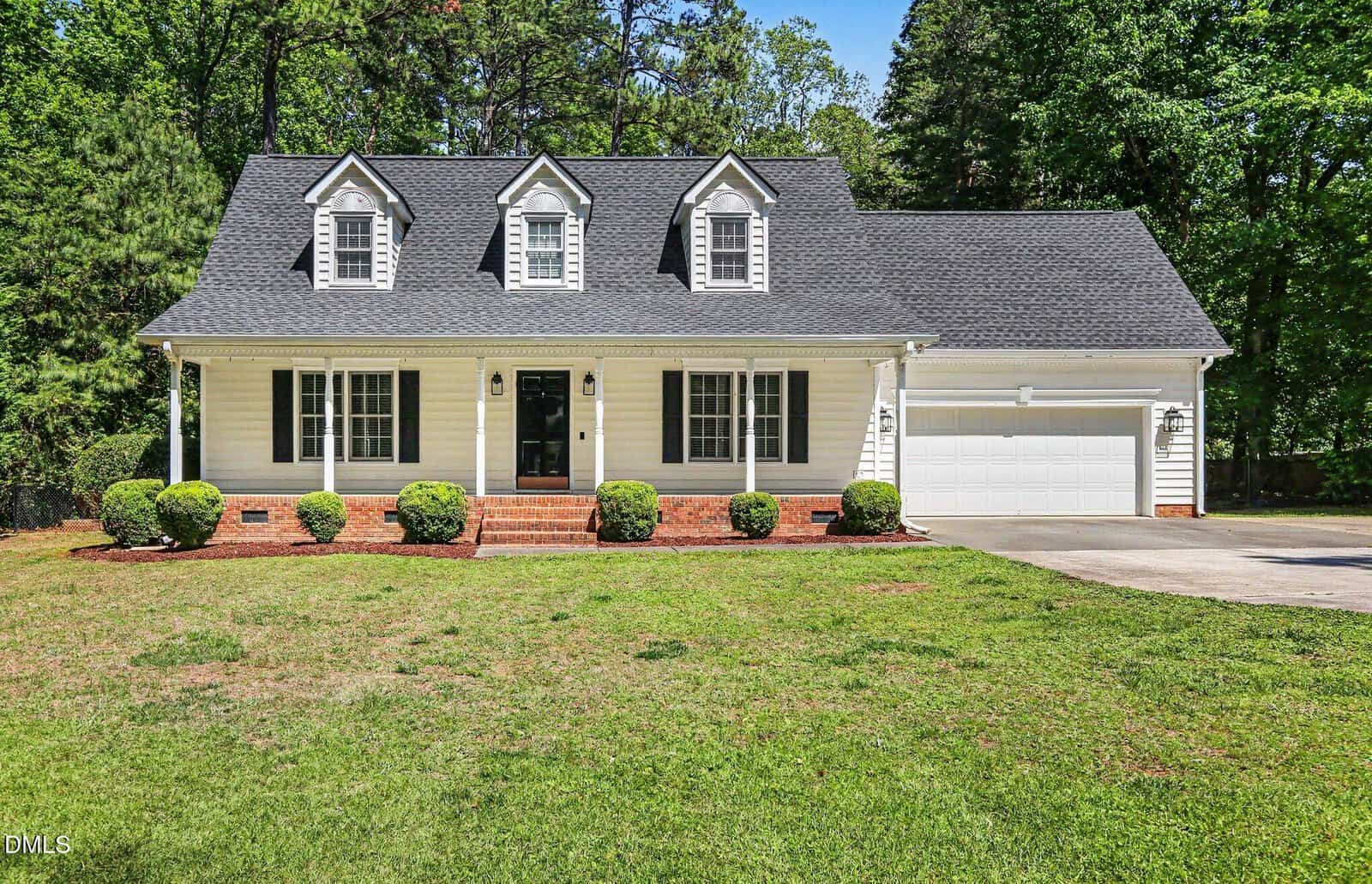 A white two-story house with dormer windows, black roof, front porch, and attached two-car garage. The house sits on a grassy lawn with shrubs and is surrounded by tall trees.