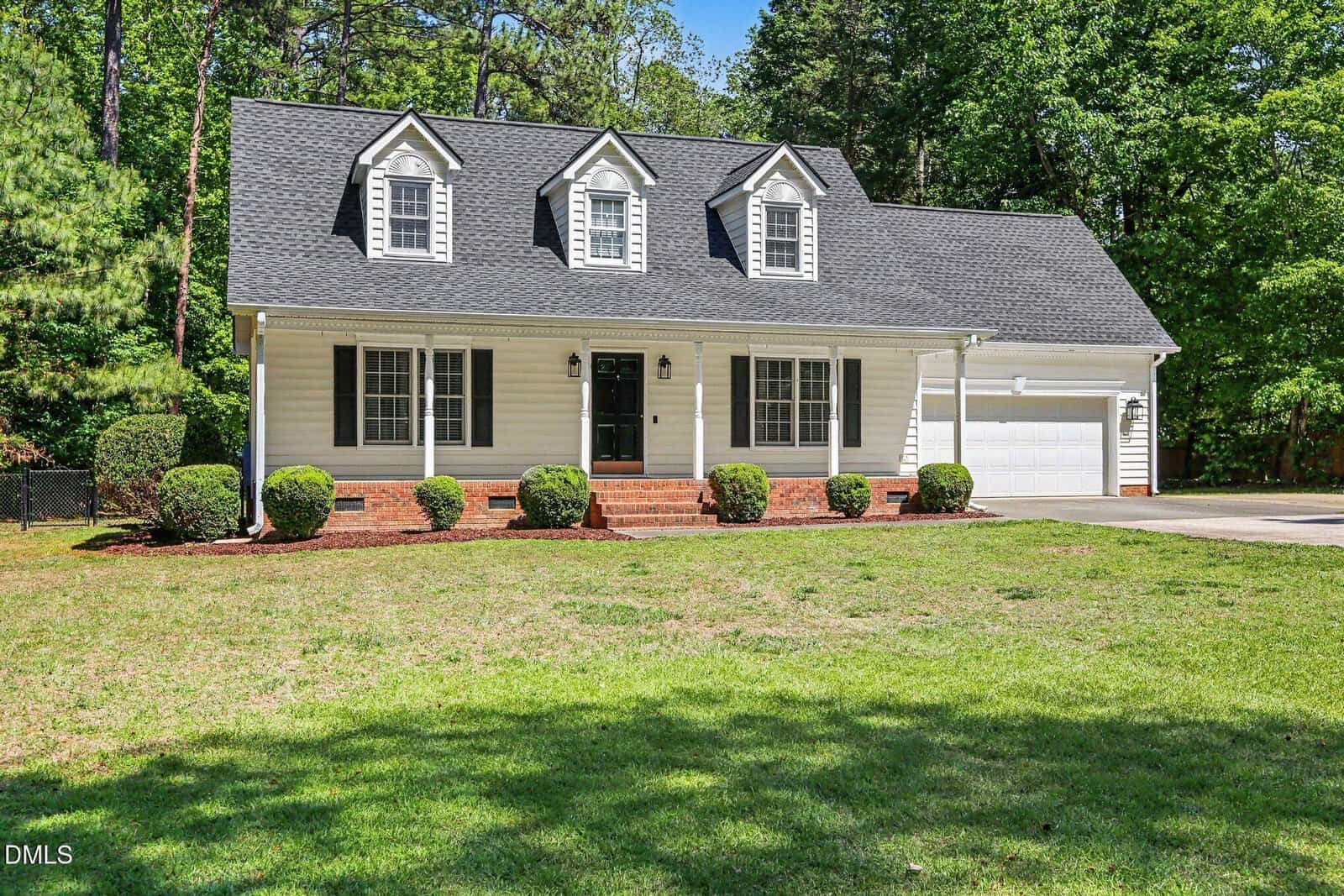 A two-story white house with black shutters, a dark gray roof, three dormer windows, and a front porch with brick steps. The house is surrounded by trimmed bushes and green grass, with trees in the background.