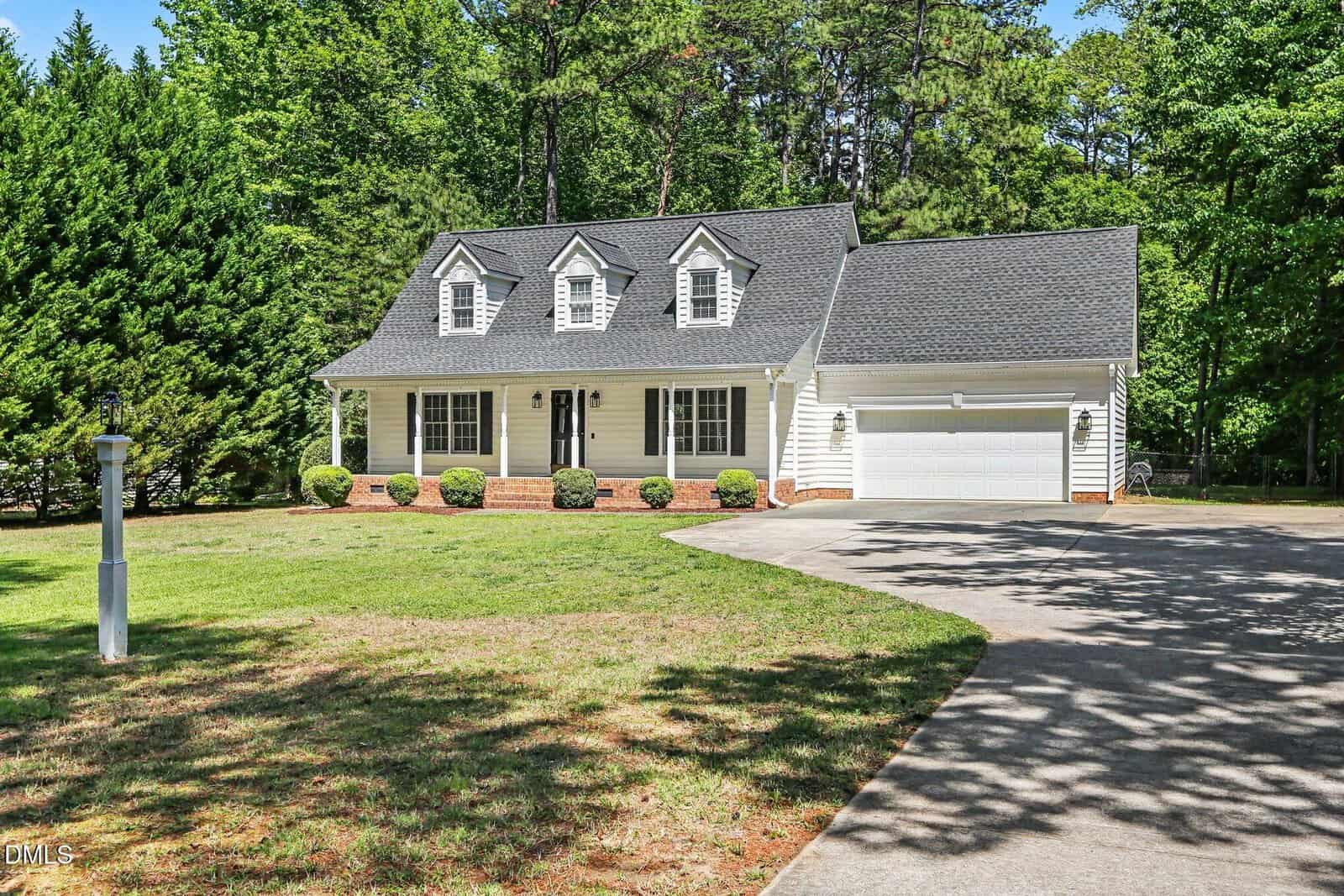 A two-story suburban house with a light yellow exterior, three dormer windows, a front porch, and an attached two-car garage; surrounded by a lawn, bushes, and tall trees in the background.