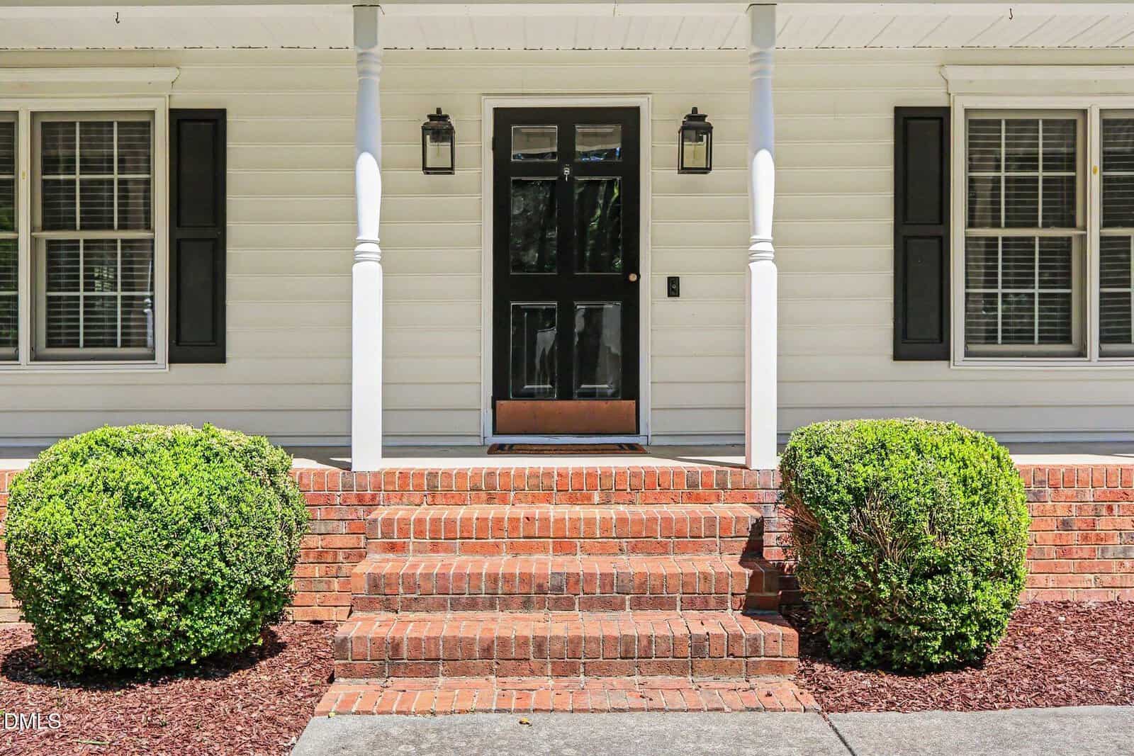 Front porch of a house with white siding, black front door with glass panels, brick steps, two white columns, two lantern lights, and two neatly trimmed bushes on either side.