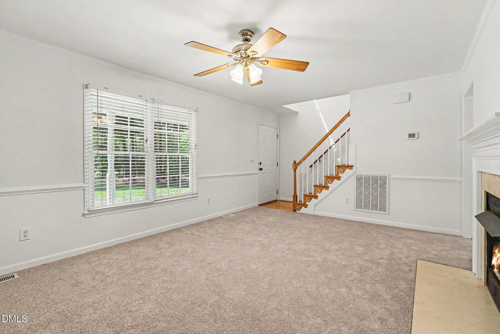 A bright living room with beige carpet, white walls, a ceiling fan, large window, fireplace on the right, and a wooden staircase near the back door.