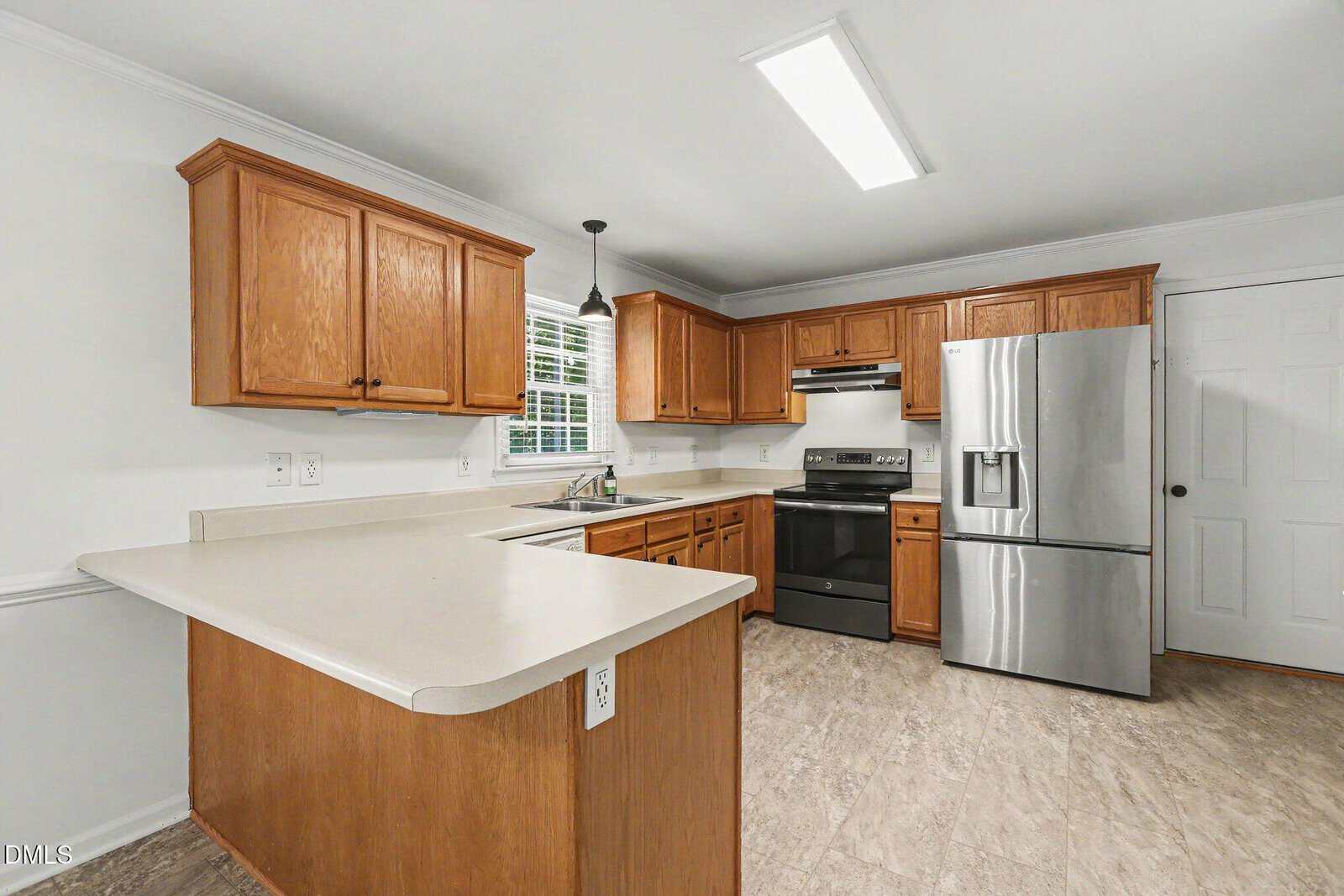 Modern kitchen with wooden cabinets, white countertops, stainless steel appliances, electric stove, double sink under a window, pendant light, and light-colored tile flooring. White walls and a white door are also visible.