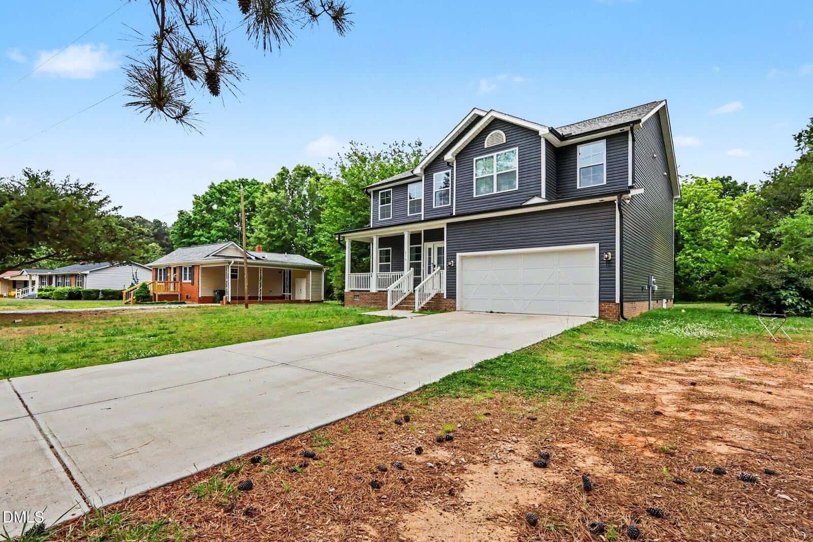 A two-story gray house with white trim, large windows, and a double garage sits beside a wide concrete driveway, surrounded by grass, trees, and neighboring homes under a clear blue sky.