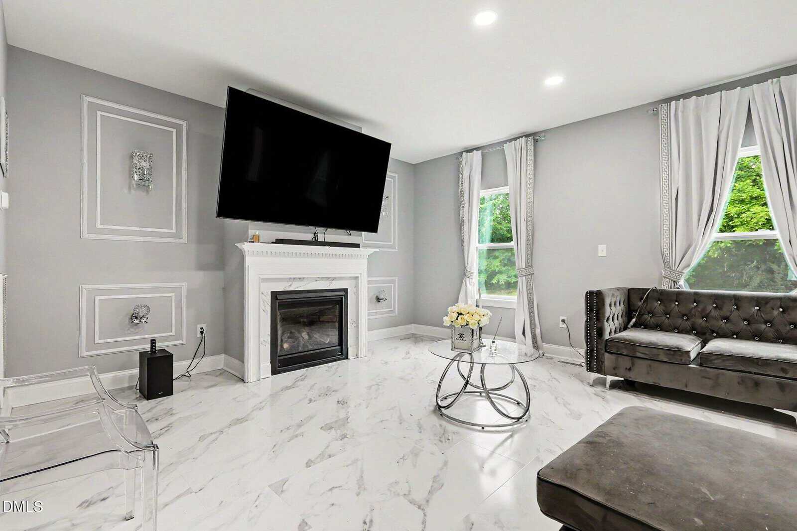 Modern living room with marble tile floor, gray walls, a wall-mounted TV above a white fireplace, silver-gray tufted sofa, glass coffee table with white flowers, and large windows with light gray curtains.