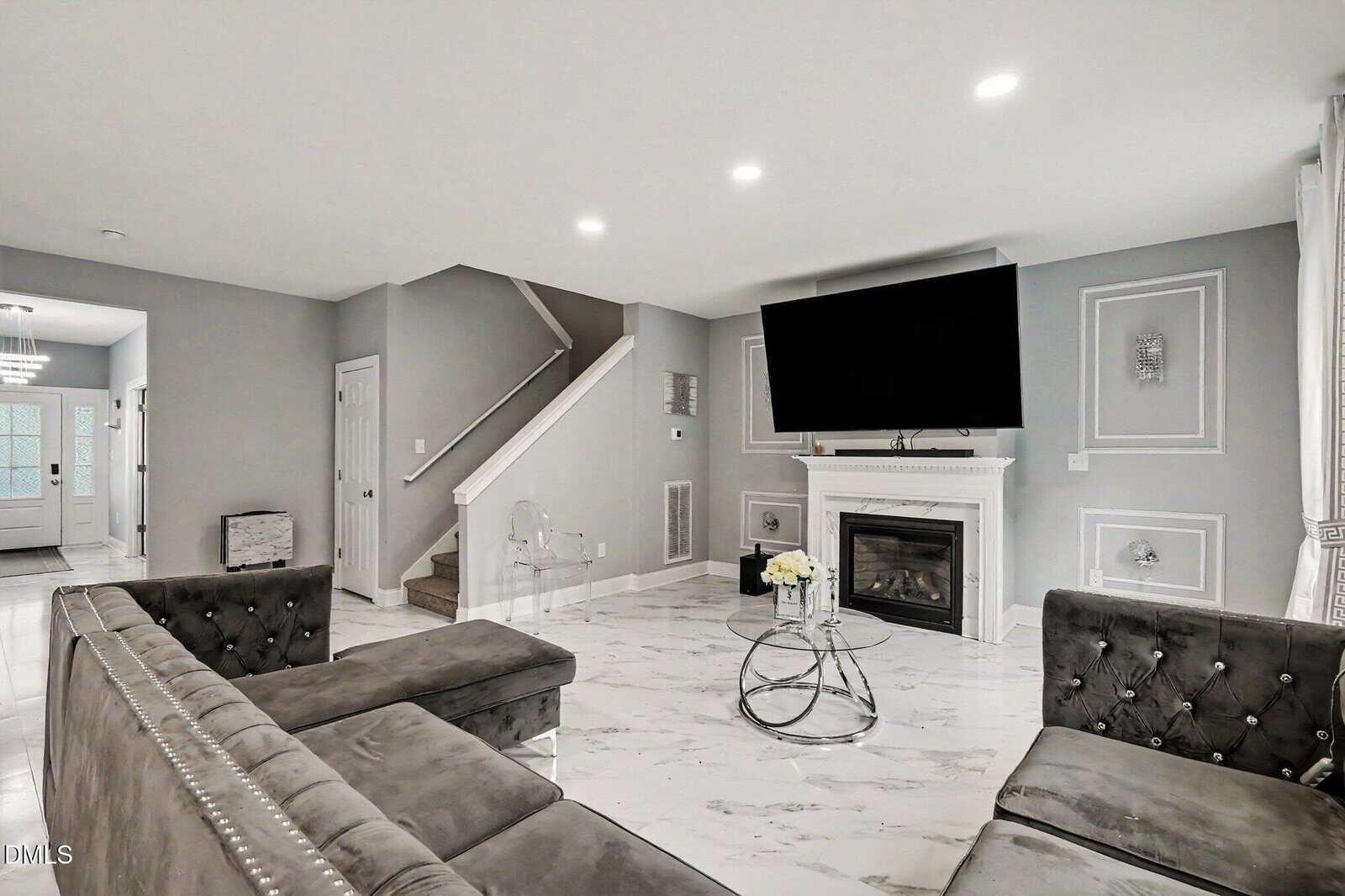 Modern living room with gray tufted sofas, marble floor, a white fireplace with a large TV above it, and a glass coffee table with flowers. Stairs and stylish wall accents are visible; the space is well-lit and contemporary.