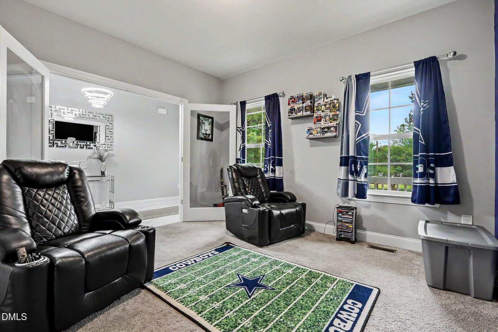 A home theater room with two black leather recliners, a Cowboys-themed rug, navy blue curtains, and shelves with collectibles. Sunlight streams through the window, highlighting the sports-themed decor.