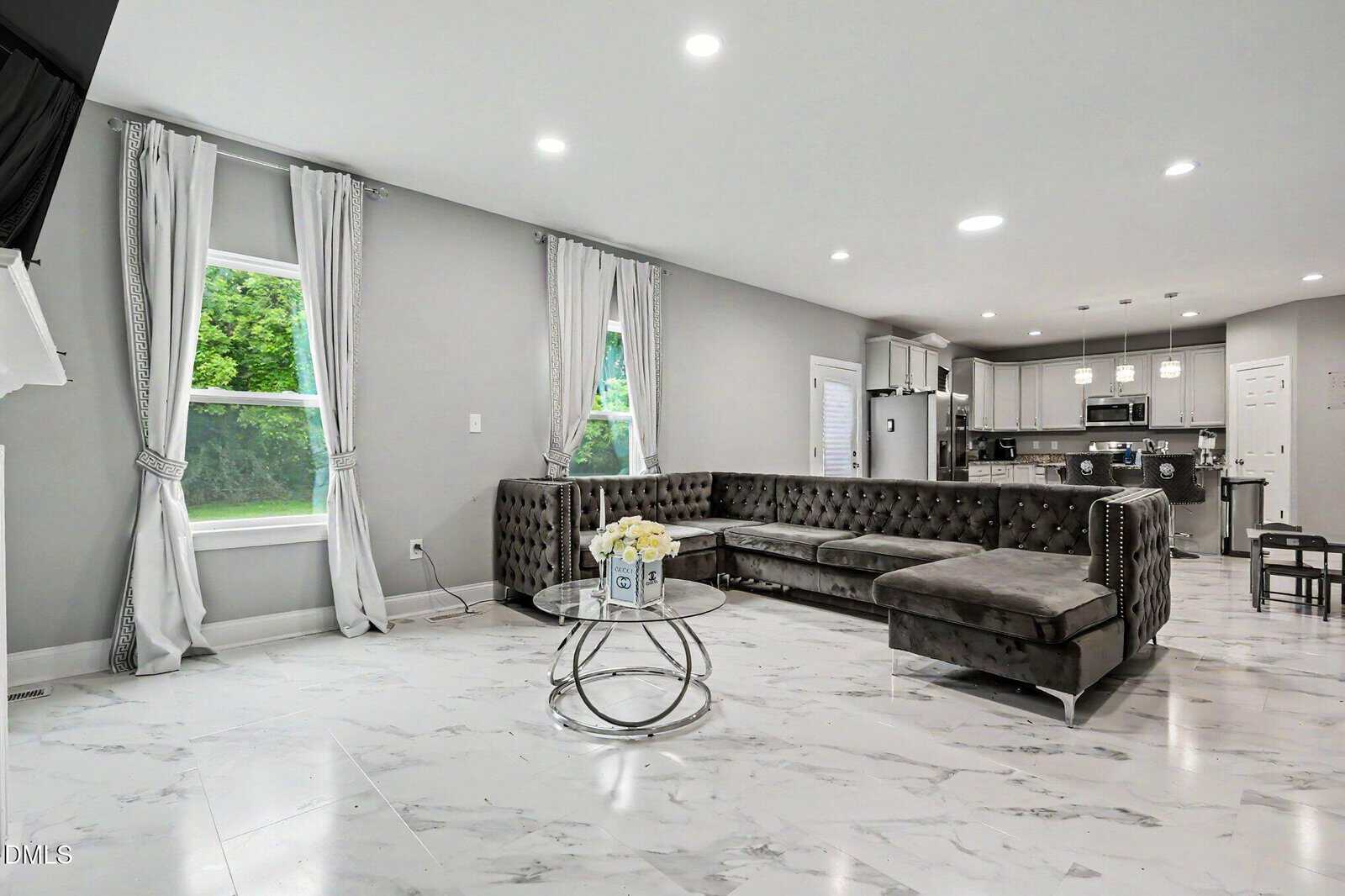Modern living room with marble floor, gray sectional sofa, round glass coffee table with flowers, large windows with curtains, and an open kitchen in the background. Bright, spacious, and stylish interior design.