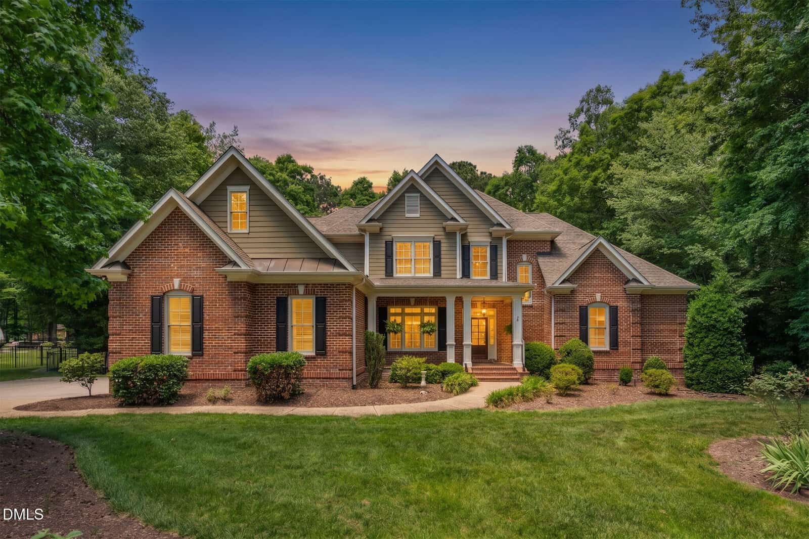 A two-story brick house with multiple peaked roofs, large windows, and a covered front porch, surrounded by neatly trimmed bushes and trees, at sunset. A green lawn extends across the front yard.