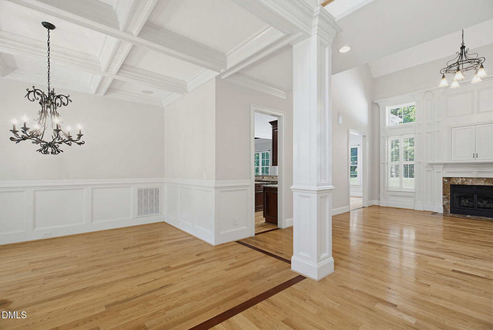 Bright, empty room with hardwood floors, coffered ceilings, white walls, two chandeliers, fireplace, tall windows, and an open doorway leading to a kitchen area.