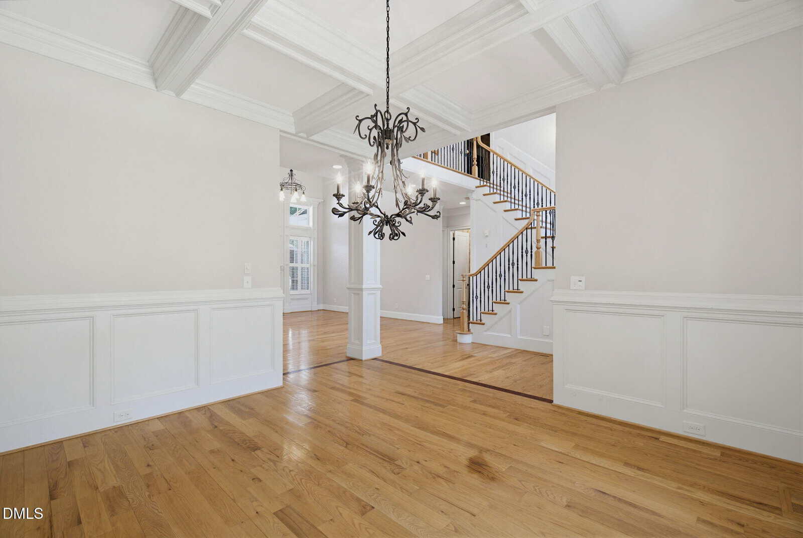 Bright dining room with coffered ceiling, ornate chandelier, light hardwood floors, white wainscoting, and an open view to a hallway with a staircase and more natural light.