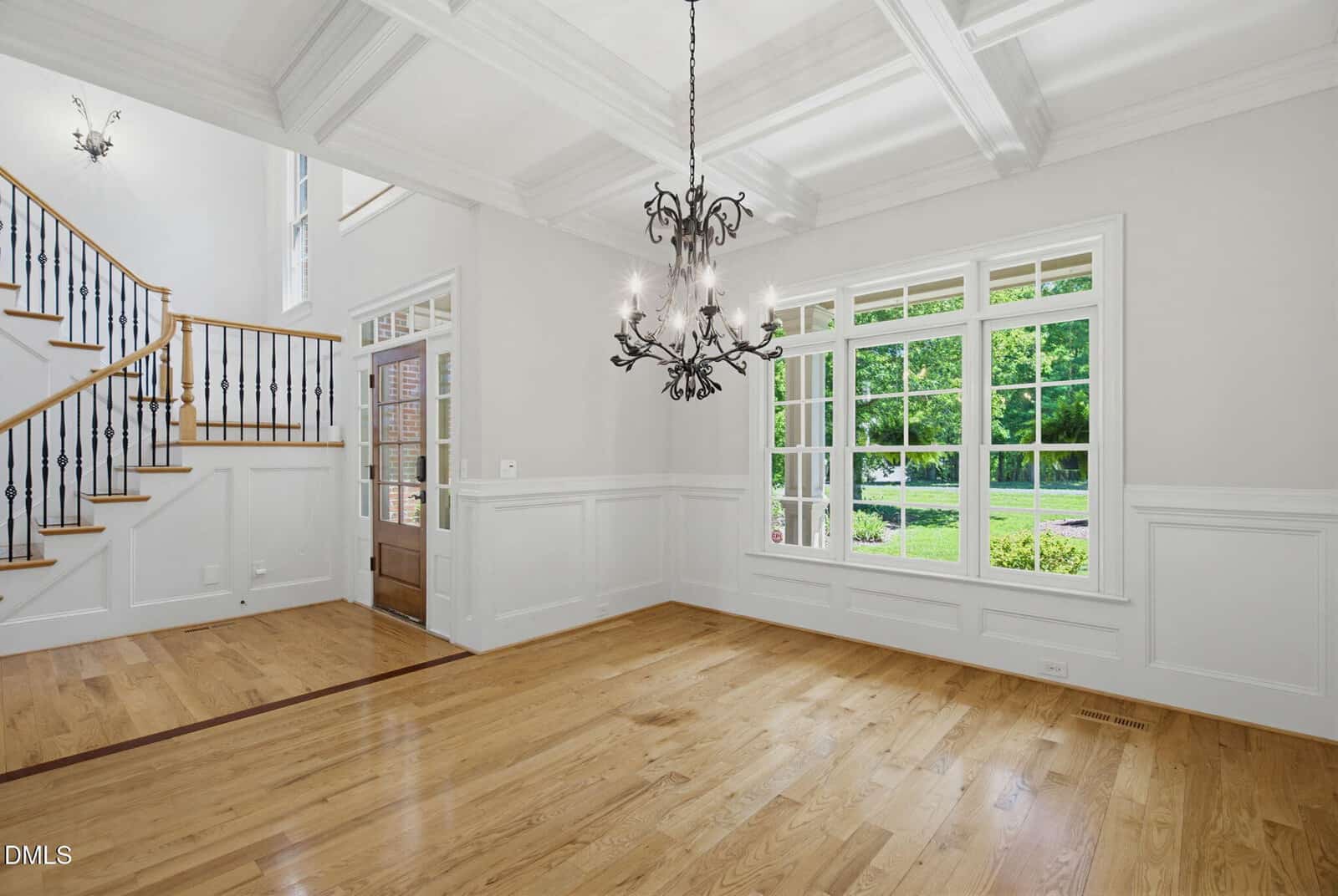 Bright empty room with hardwood floors, large windows, white coffered ceiling, ornate chandelier, white walls with wainscoting, and a staircase with wooden steps and black balusters.