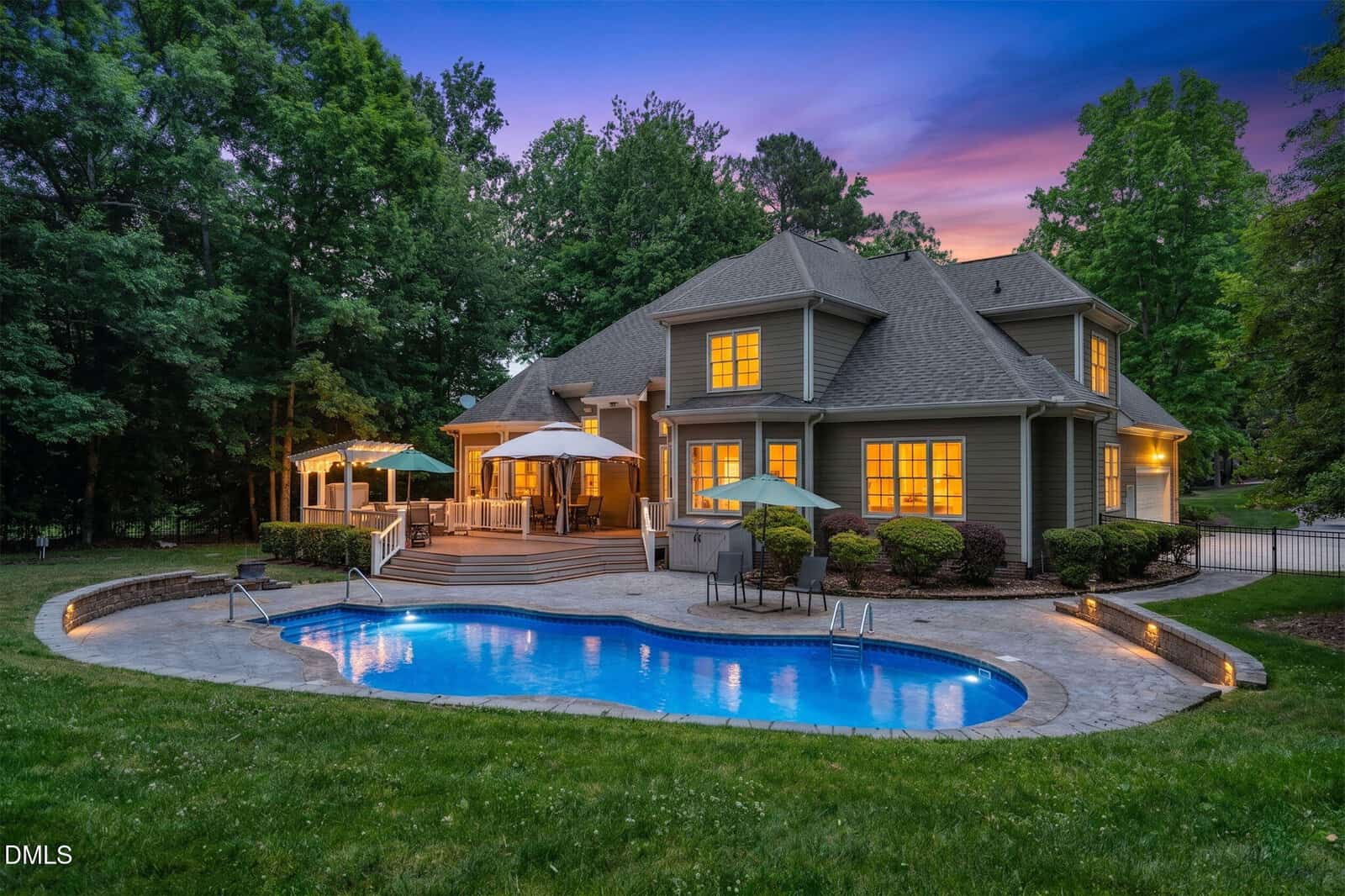 A spacious house with large windows and outdoor lights sits beside a curving swimming pool, surrounded by a stone patio, lounge chairs, and umbrellas, with trees and a vibrant sunset sky in the background.