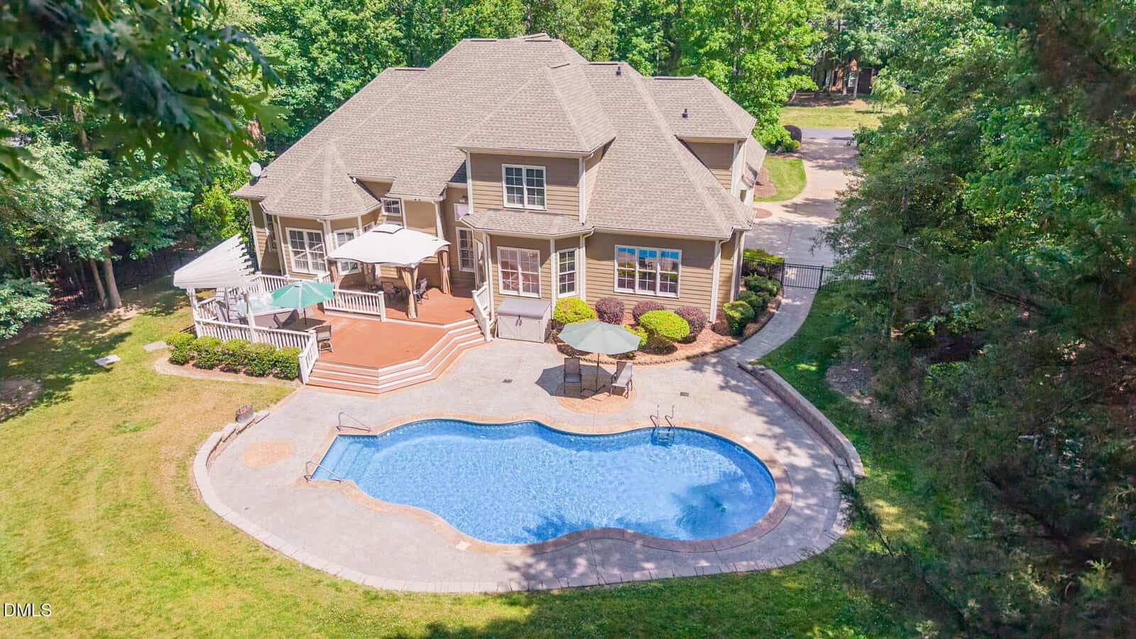 Aerial view of a large beige house with multiple roof peaks, a spacious wooden deck with umbrellas, and a curved in-ground swimming pool surrounded by patio and trees in a landscaped backyard.