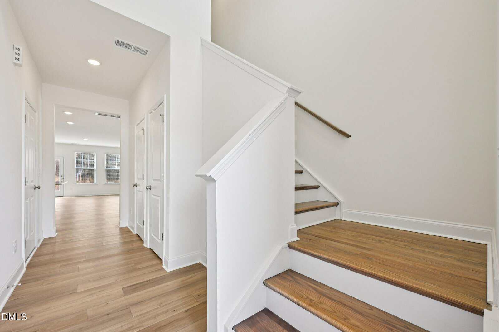 A bright, modern hallway features light wood floors, white doors, and a wooden staircase with white risers and treads.