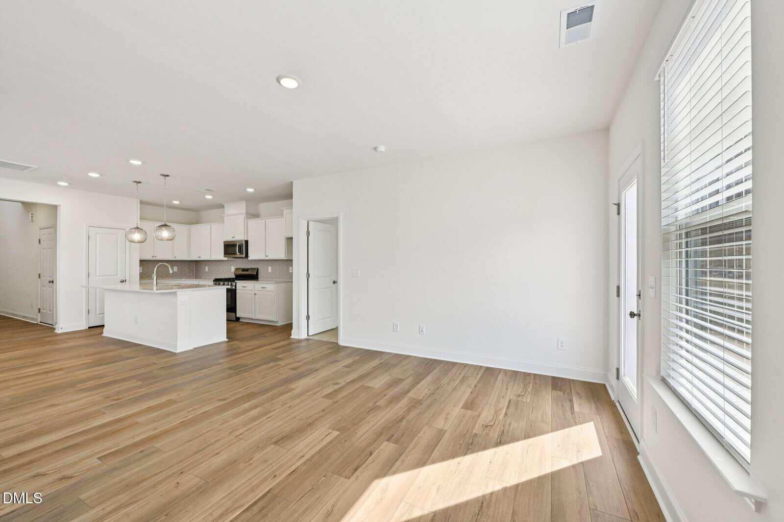 An open-concept living area with a modern kitchen, white cabinets, an island, and hardwood floors. Sunlight streams in from a large window.