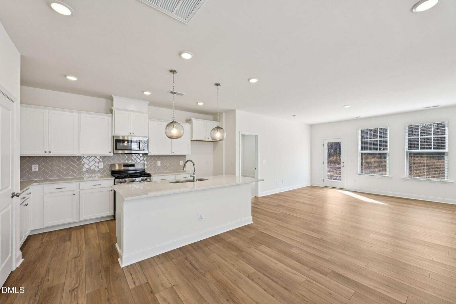 Modern, spacious kitchen with white cabinets, stainless steel appliances, pendant lights over an island, and an open living area featuring light wood floors and large windows letting in natural light.