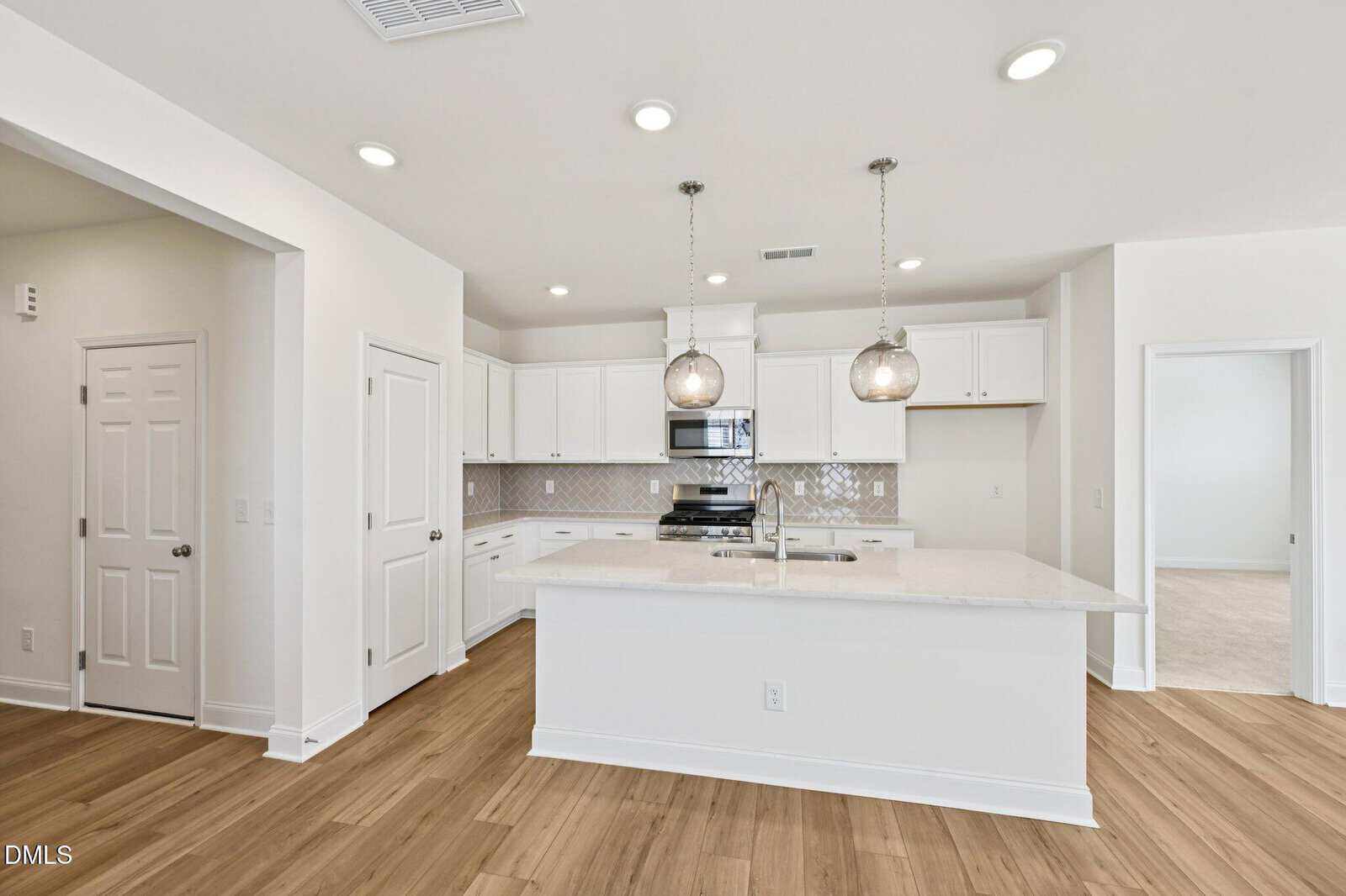 Modern kitchen with white cabinets, stainless steel appliances, two pendant lights over a large island with sink, light wood flooring, and a neutral backsplash. A pantry and open doorway are visible in the background.
