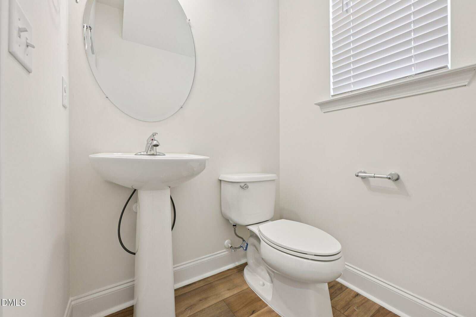 A white pedestal sink, toilet, and oval mirror in a powder room with light wood floors and a window with white blinds.