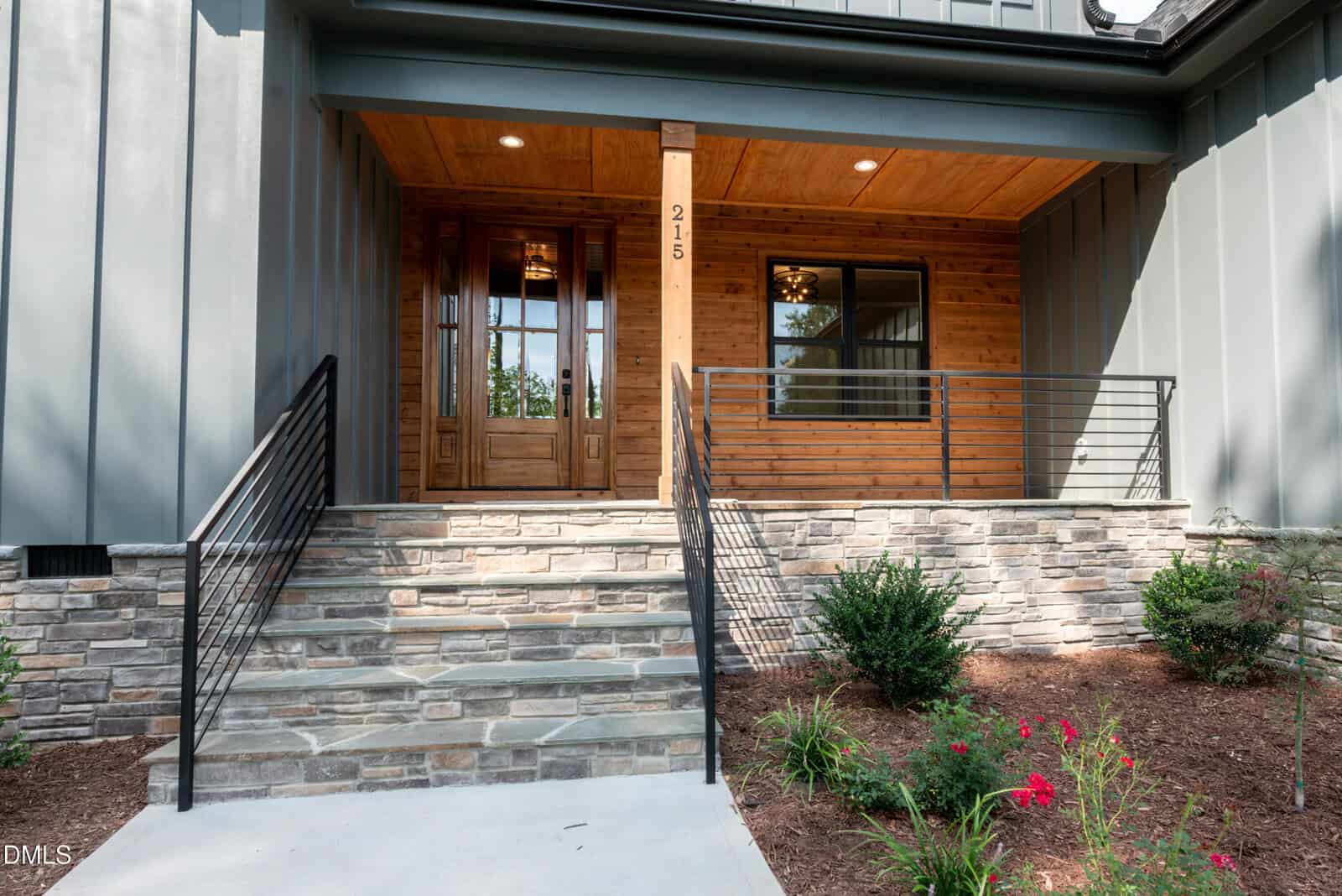 A modern house entrance with stone steps, black metal railings, a wooden front door, and a covered porch with wood paneling, surrounded by landscaped bushes and flowers.